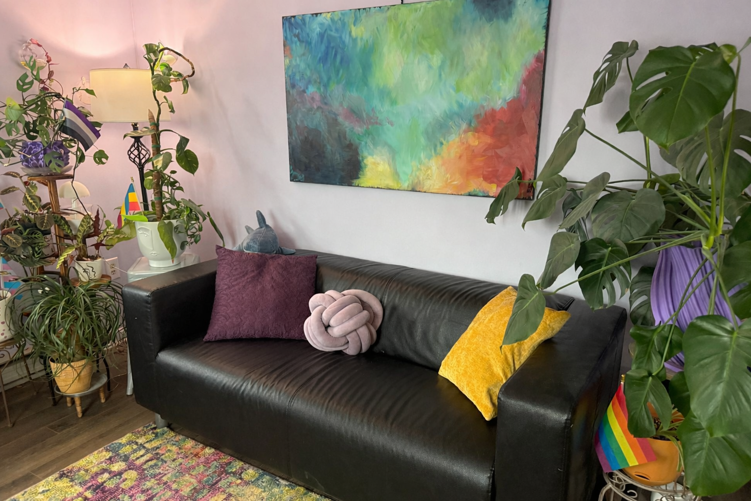 Living room with a black leather sofa decorated with colorful pillows, surrounded by various houseplants, including a large monstera on the right, a plant with purple leaves, and a decorative lamp on a side table. A colorful abstract painting hangs on the wall.