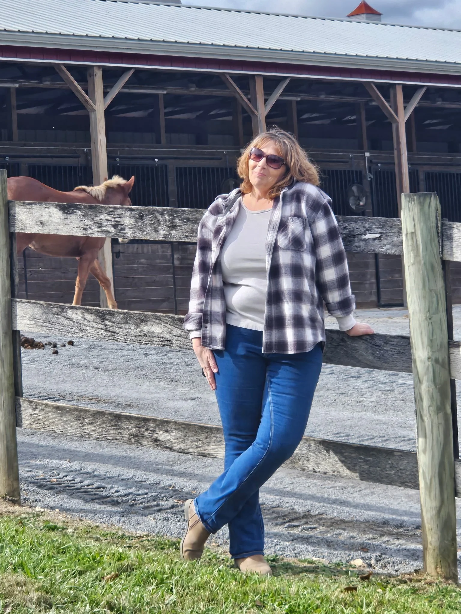 A woman wearing sunglasses, a plaid shirt, a light-colored top, and blue jeans leaning against a wooden fence at a farm, with a horse behind her and a barn in the background.