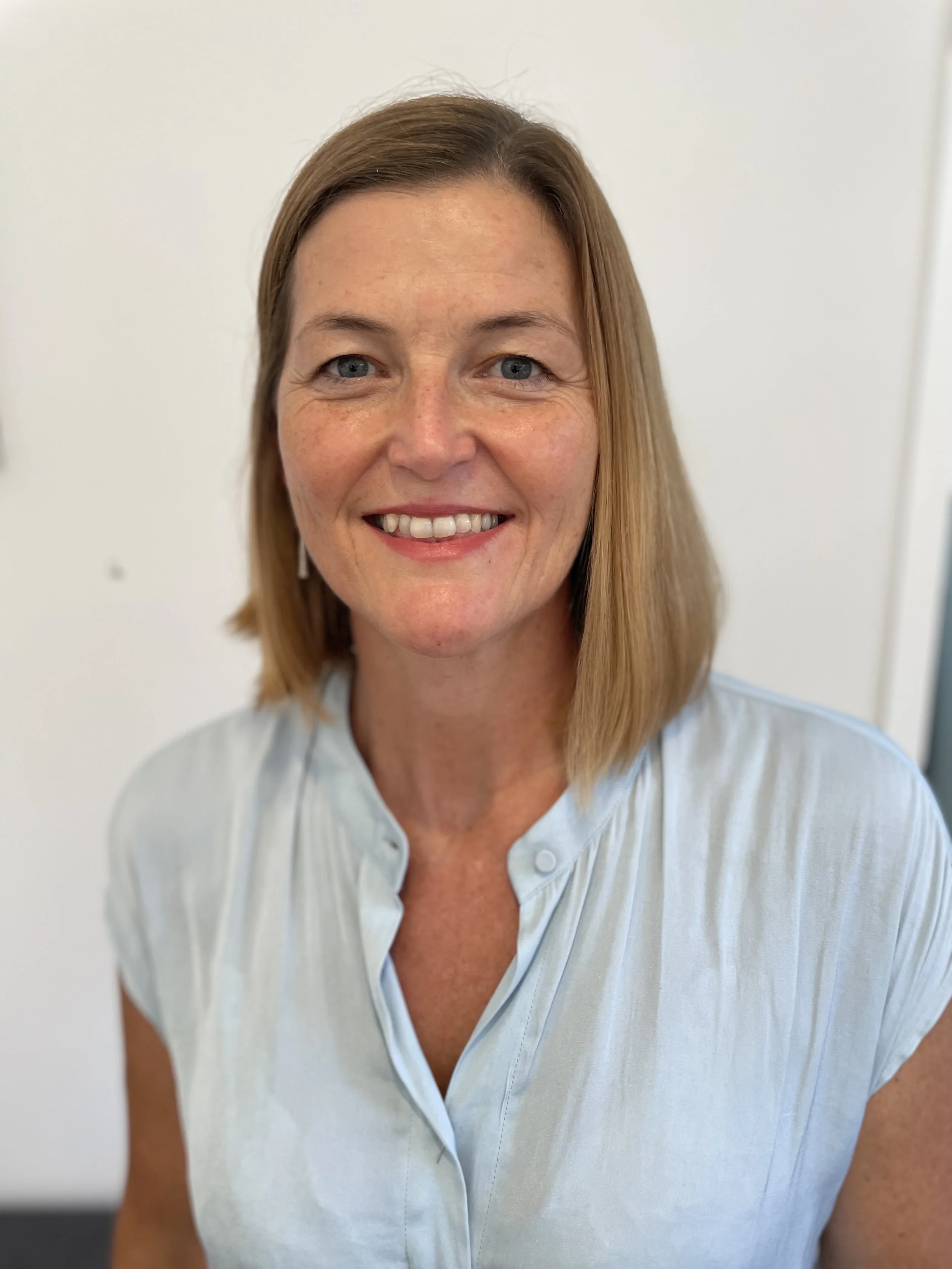A woman with shoulder-length blonde hair and blue eyes, smiling, wearing a light blue blouse, standing in front of a plain off-white wall.