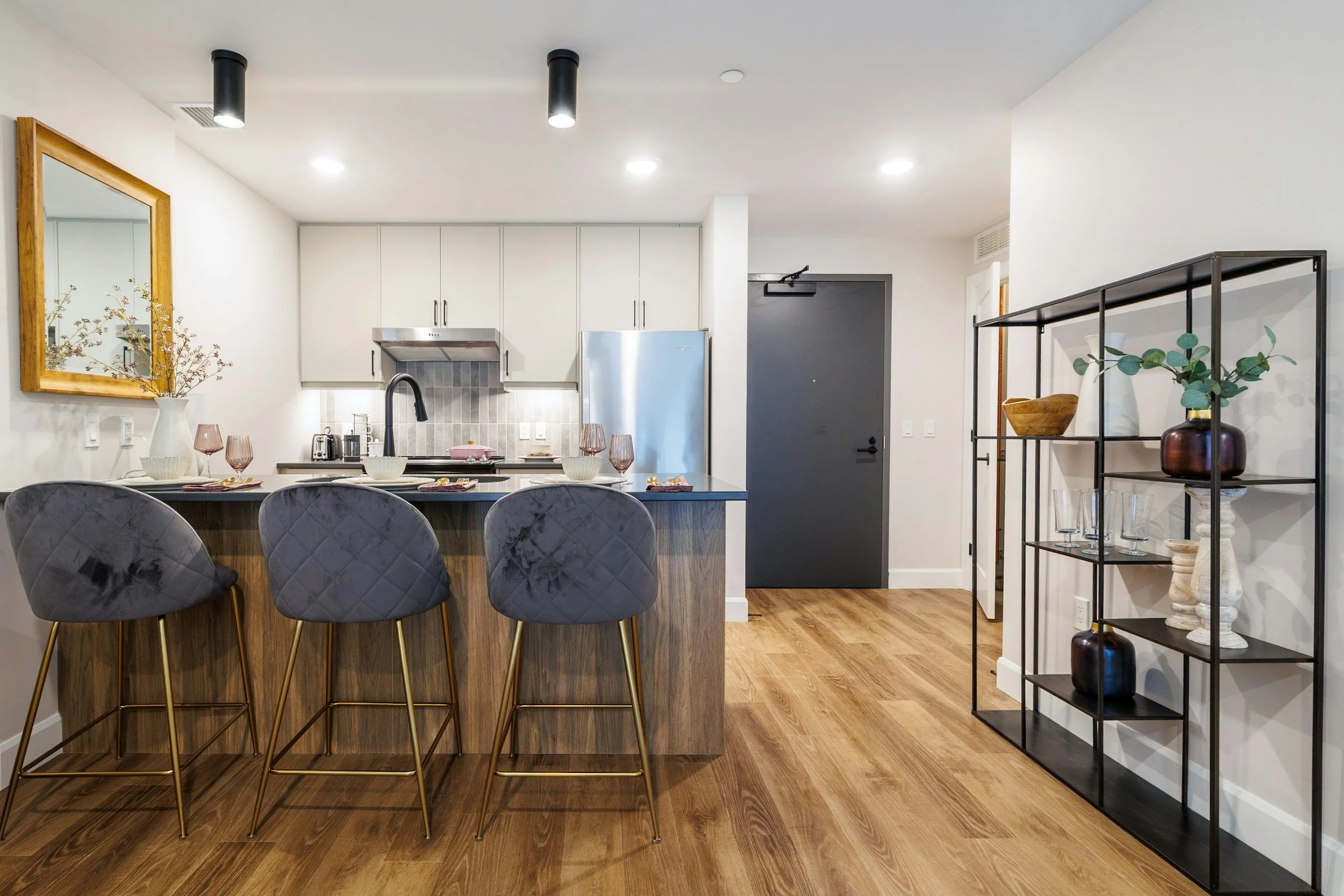 Modern kitchen with a black countertop, grey bar stools, white cabinets, stainless steel refrigerator, black door, and a black metal shelving unit with decorative vases and greenery on a hardwood floor.