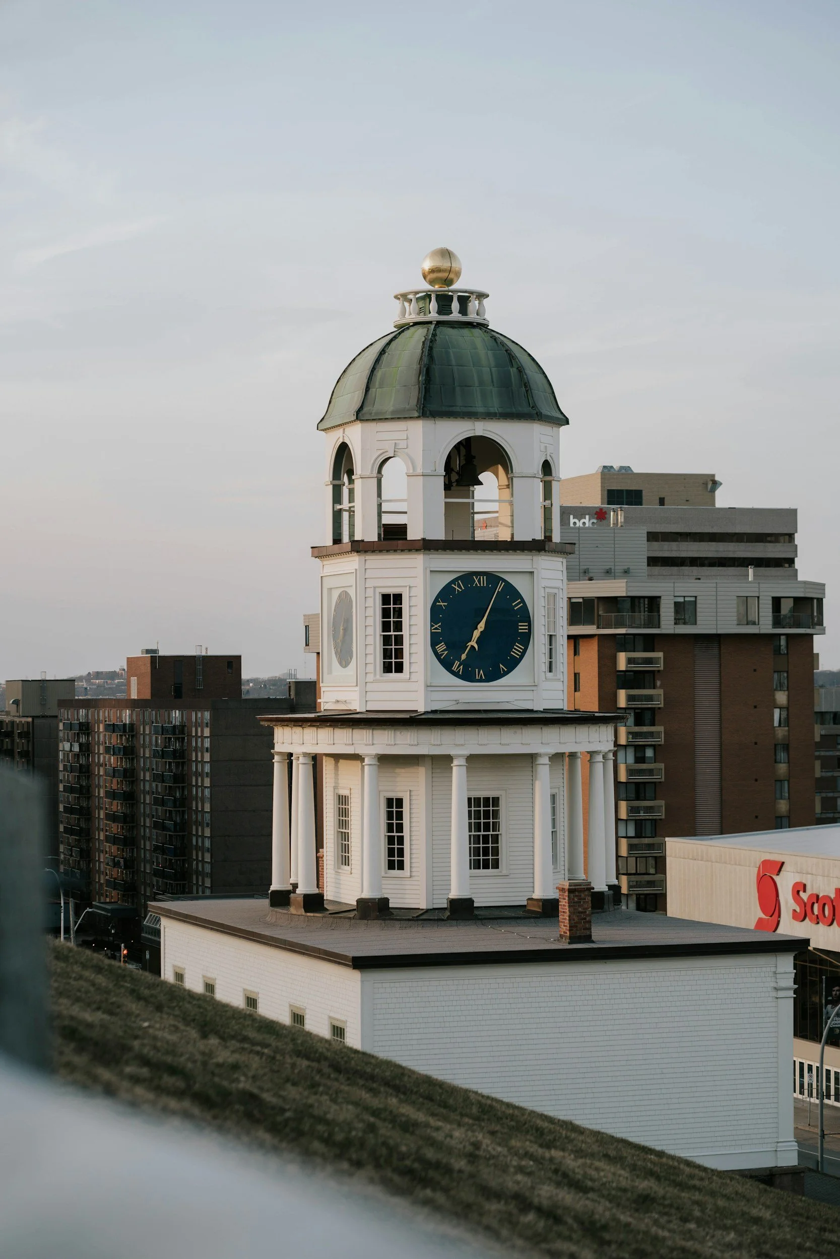 A historic white clock tower in Halifax with a green dome and gold ornament on top, showing the time as approximately 6:02, with modern buildings in the background.
