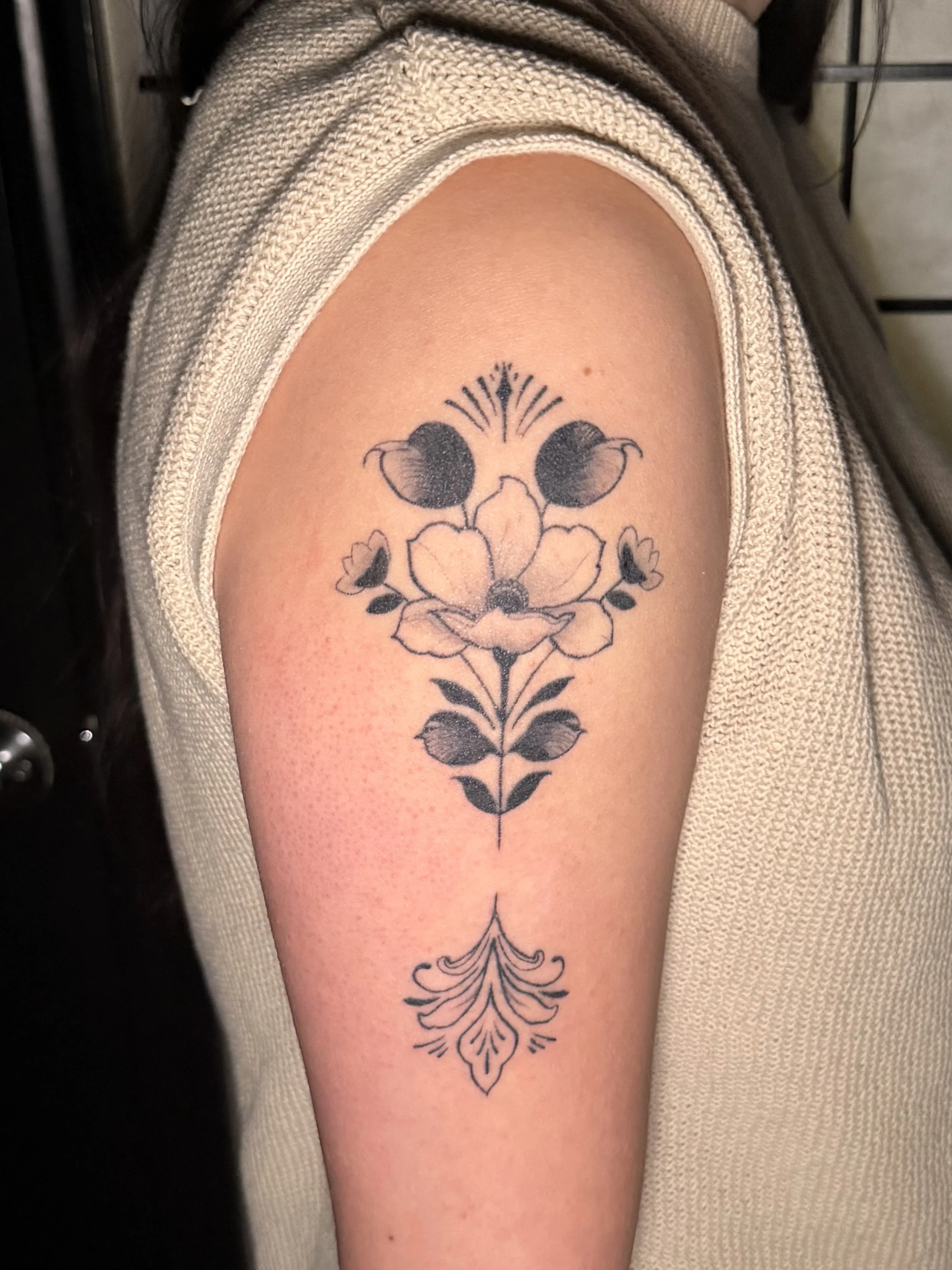 A tattoo on a person's upper arm featuring a symmetrical floral design with leaves and decorative elements.