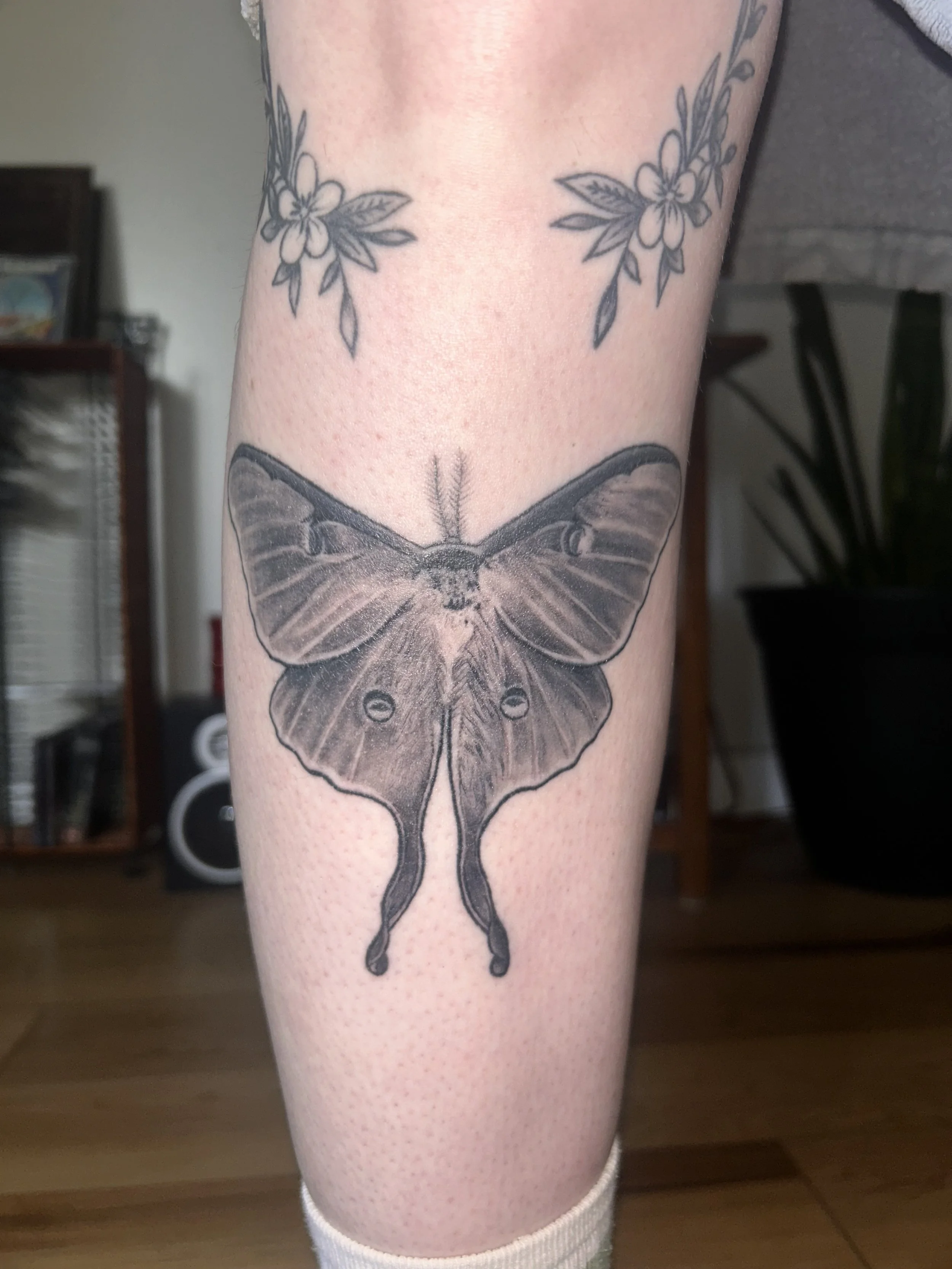 A black and gray tattoo of a moth on a person's calf, with smaller flower tattoos above it.