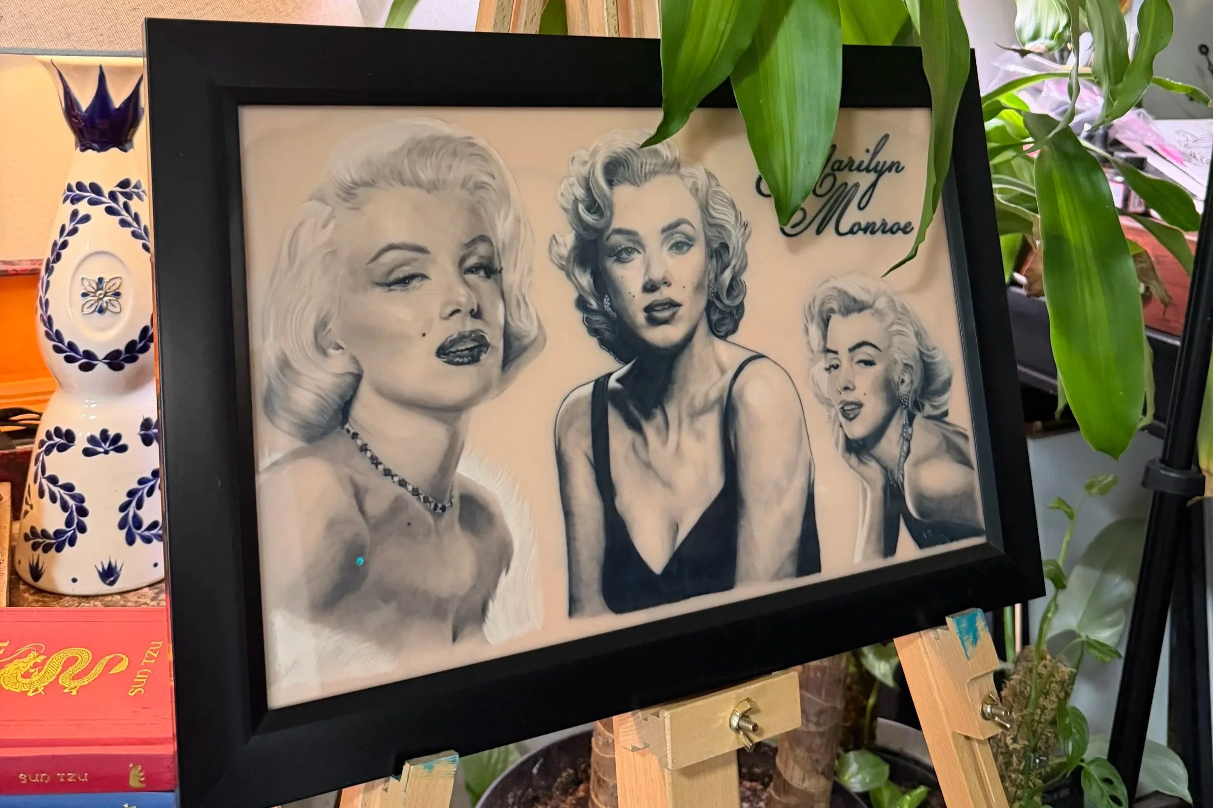 A framed black-and-white portrait of Marilyn Monroe with three different expressive images of her, surrounded by green houseplants.