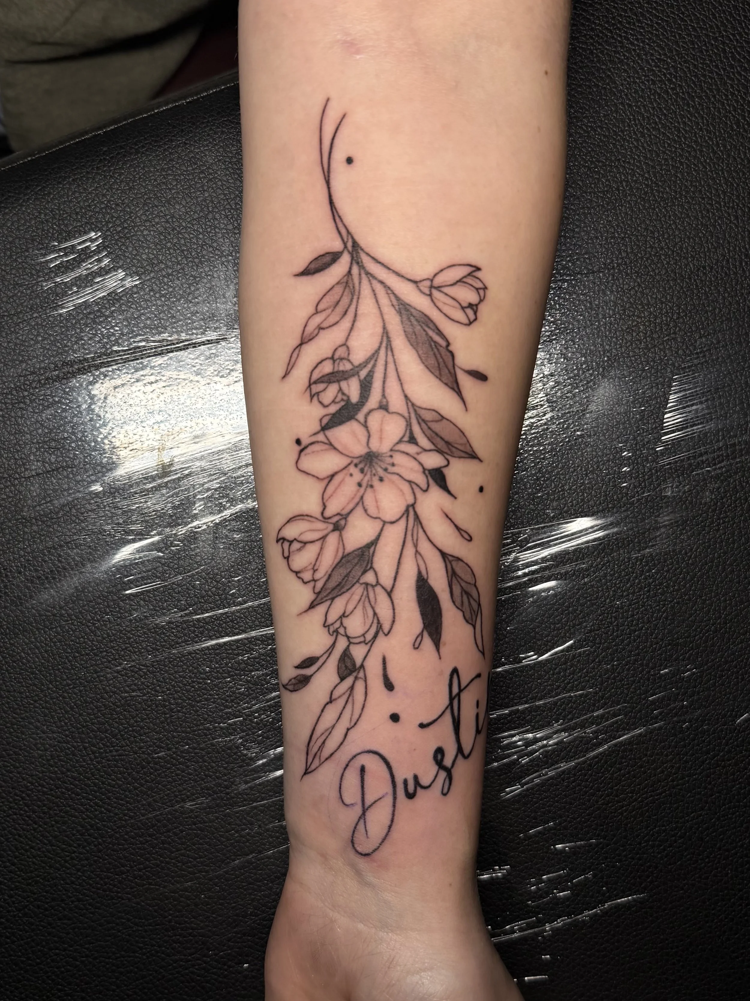 Tattoo of a floral design with the name "Dustin" written below it on a person's forearm.