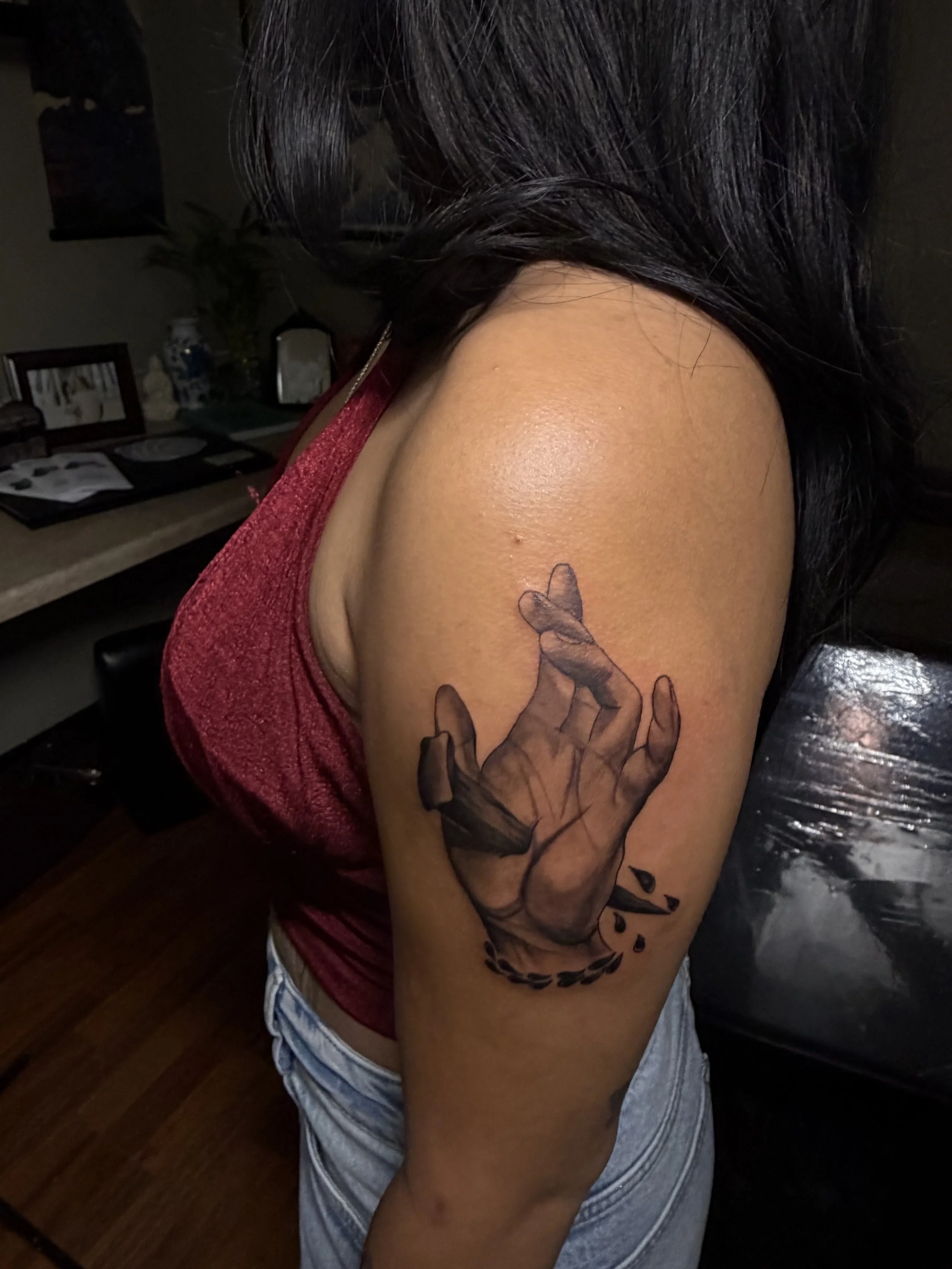 Tattoo of a hand with fingers crossed, surrounded by a small leaf and a few droplets, on a woman's upper arm.