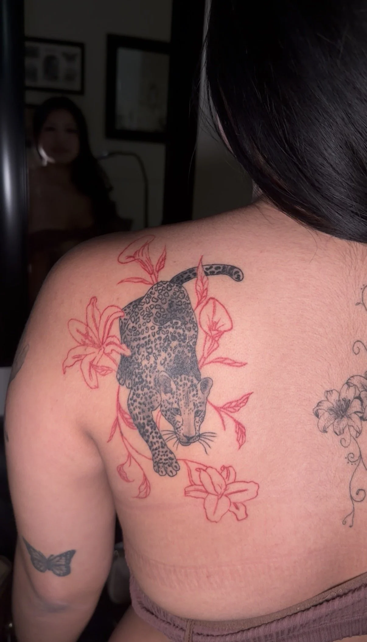 A tattoo of a leopard cub surrounded by red flowers on a person's shoulder and upper arm, with a mirror reflecting a woman in the background.