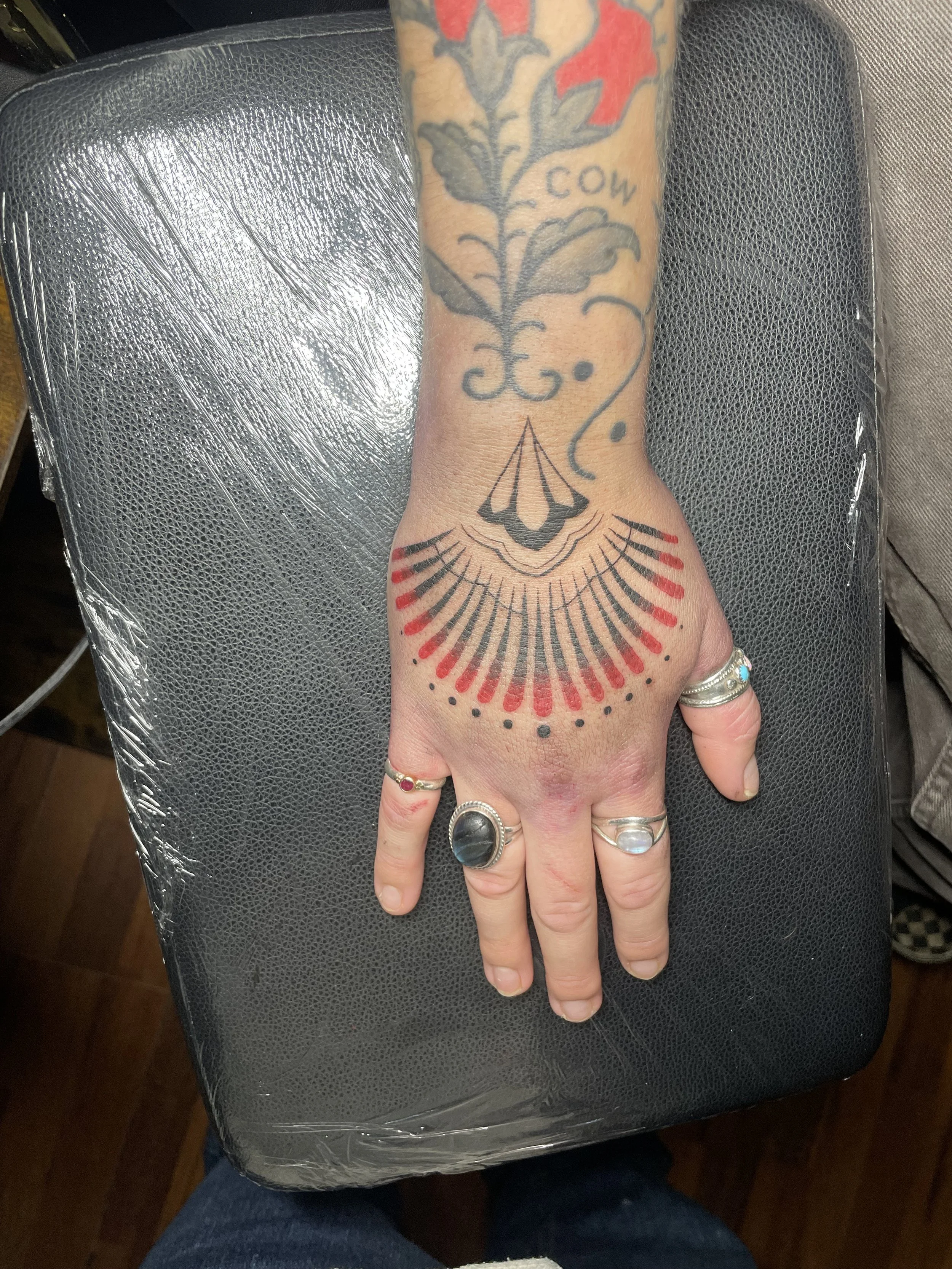 A person's hand with tattoos, including a floral design with the word 'COW' and a geometric black and red design on the wrist. The hand has multiple rings, including silver rings with stones, resting on a black textured surface covered with clear plastic wrap.