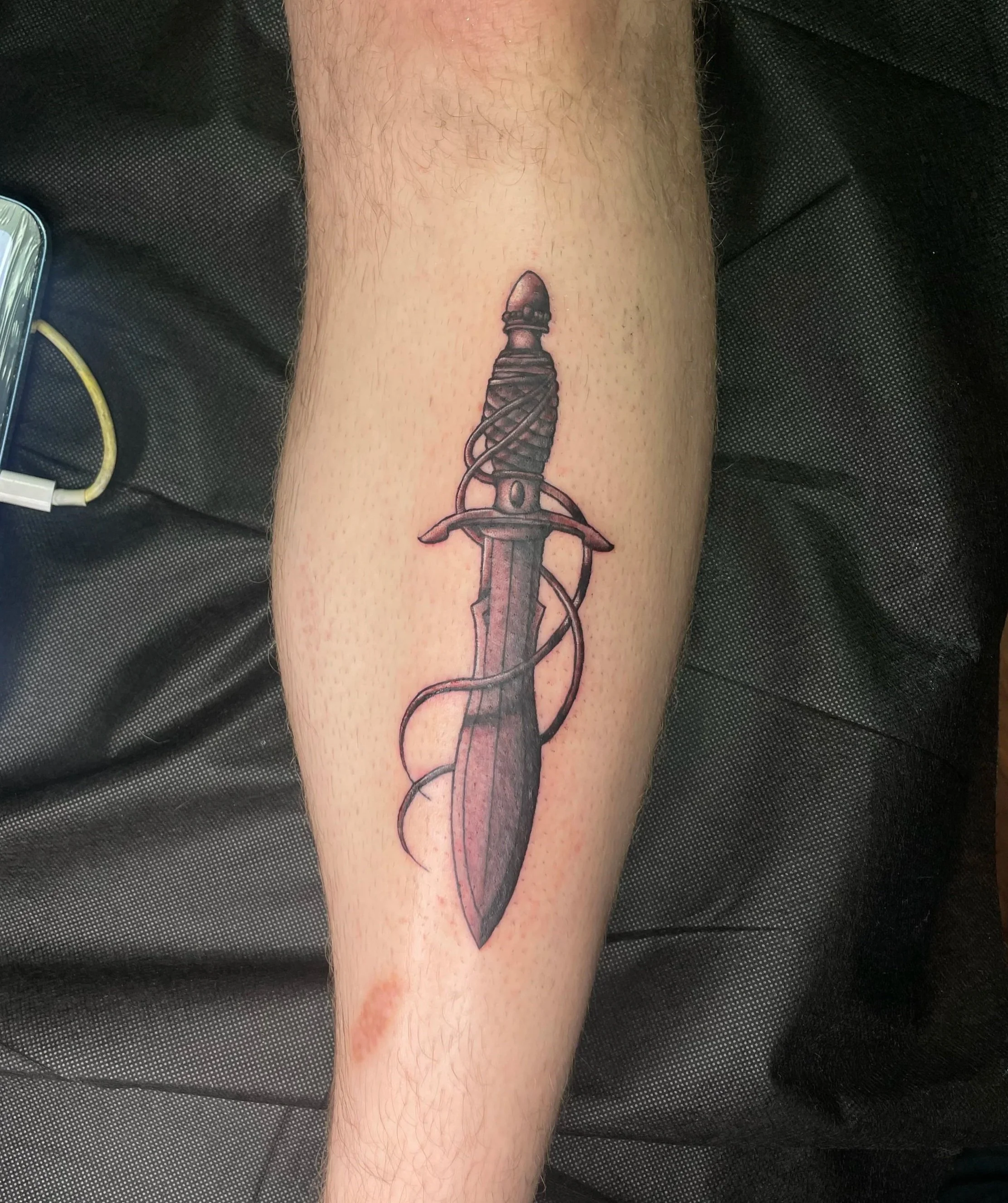 Tattoo of a dagger with a wrapped handle and a curved guard on the inner side, inked on a person's forearm.