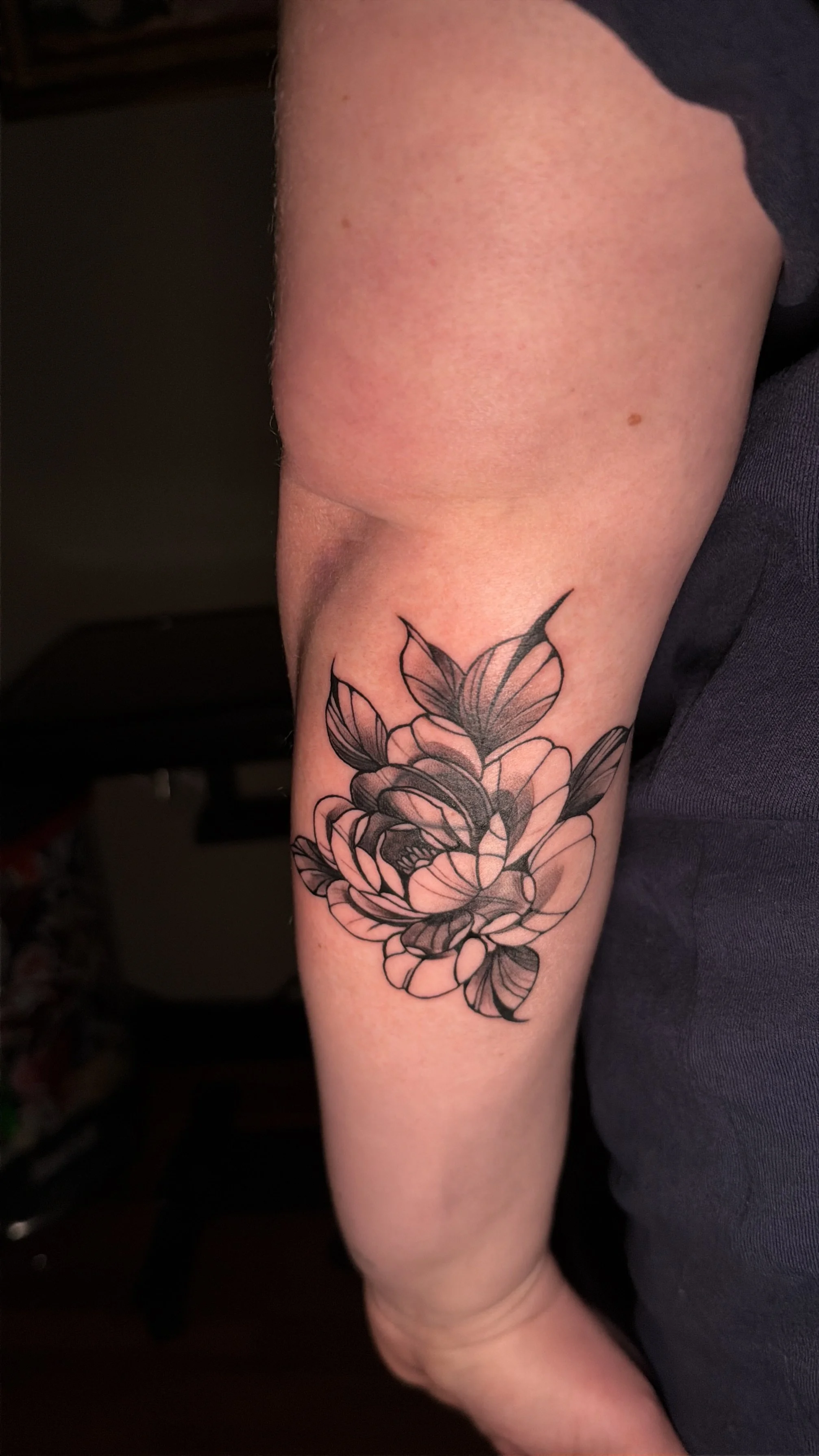A black and gray floral tattoo on a person's forearm, featuring a large blooming flower with detailed petals and leaves.