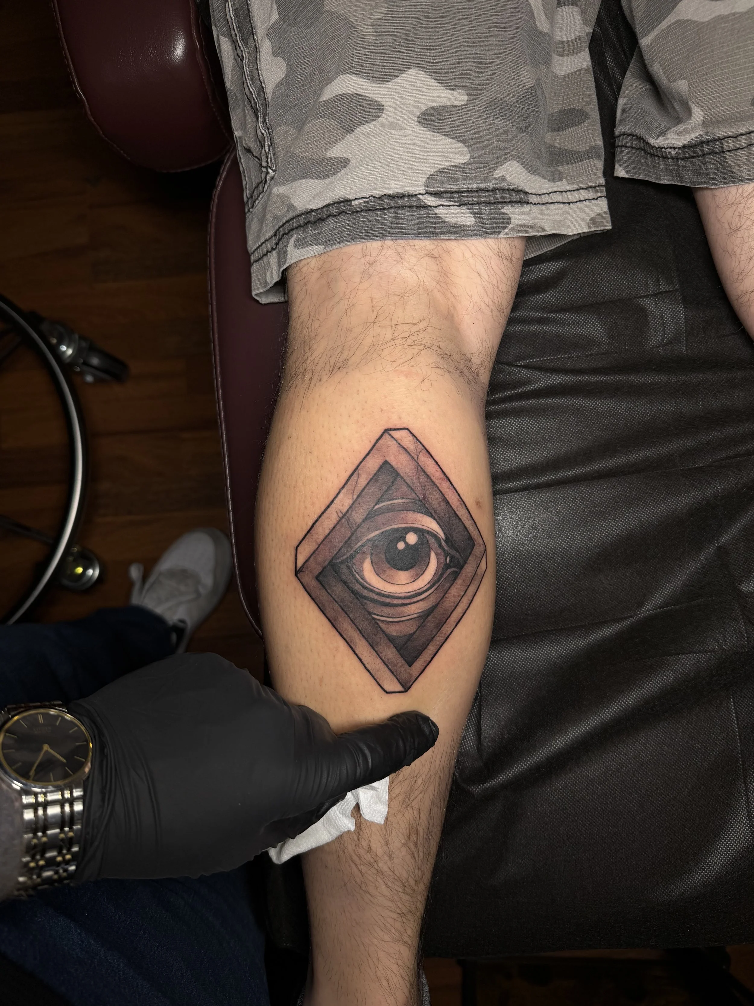 A tattoo of a realistic eye with detailed iris and eyelashes inside a 3D diamond-shaped frame on a person's lower leg.