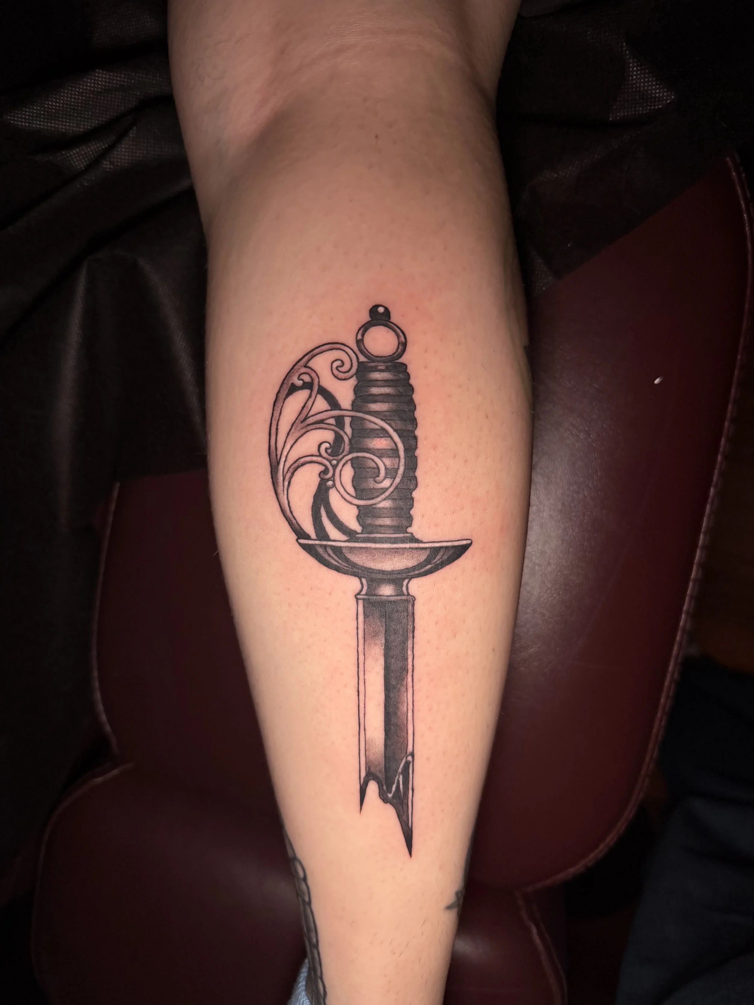 Tattoo of a sword with ornate details on the hilt, resting against a wooden surface.