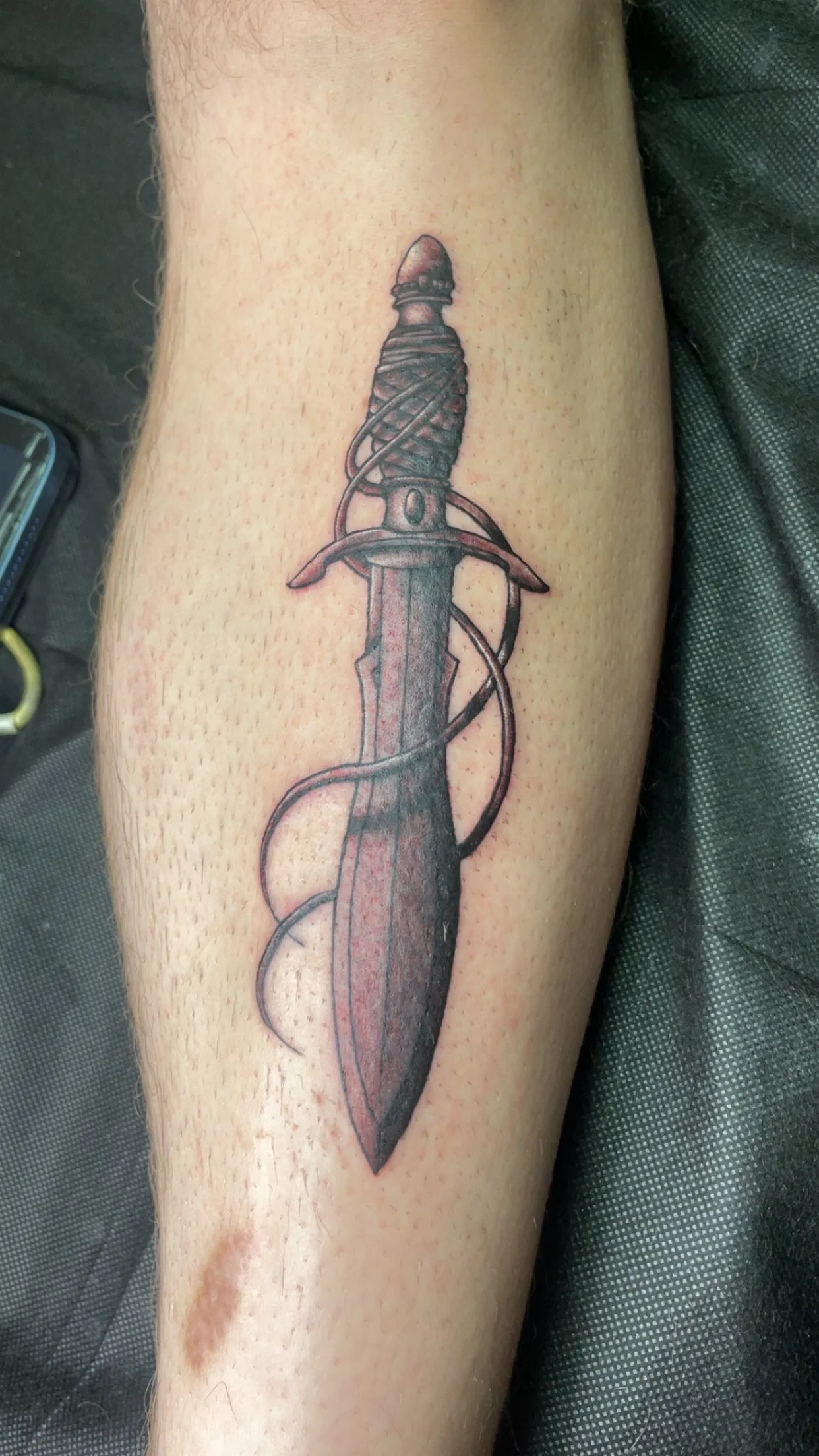 Tattoo of a dagger with a wrapped handle and a curved blade, with a cord wrapped around the dagger.