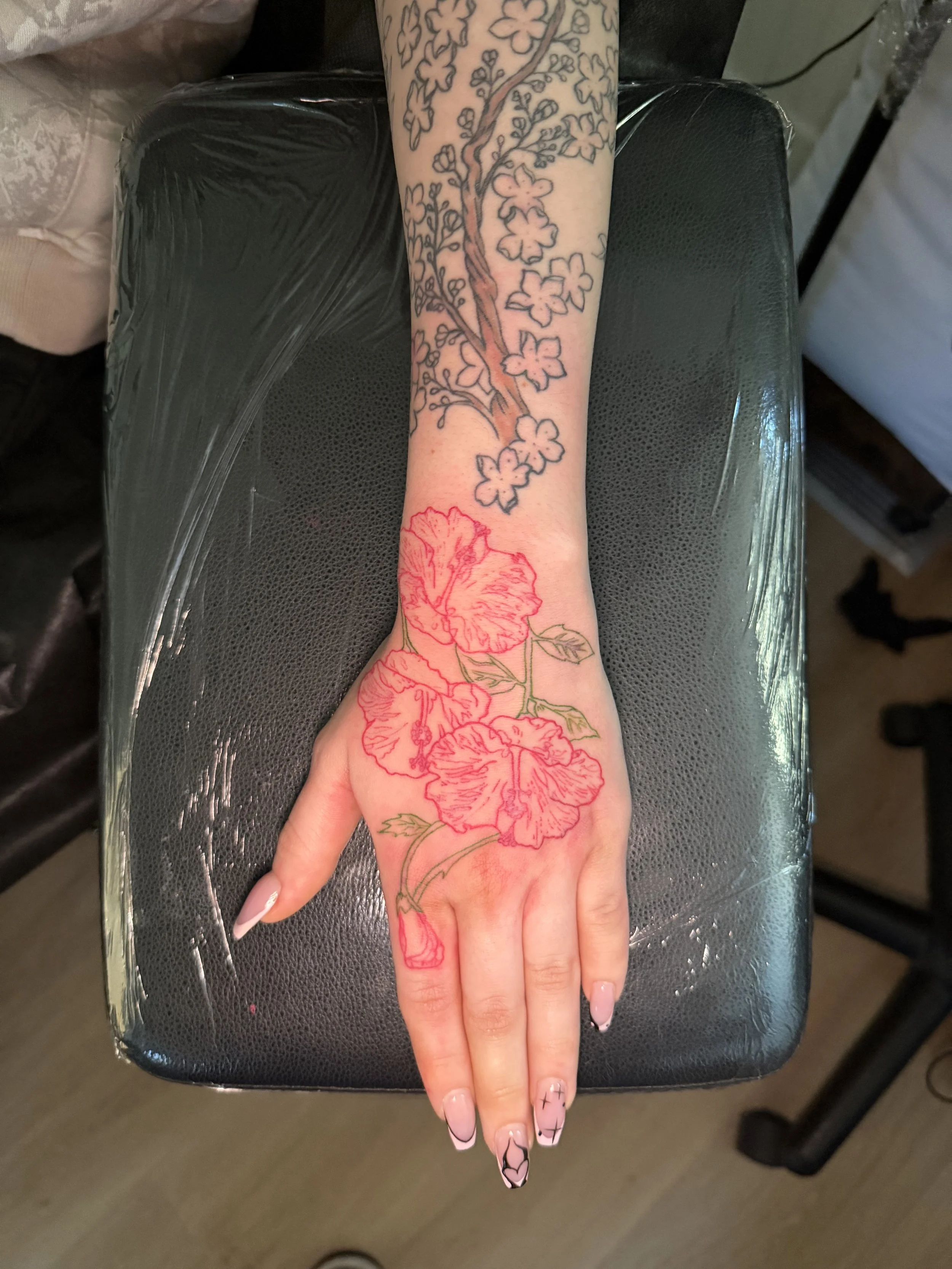 A person's arm and hand resting on a black padded surface, with a tattoo of cherry blossoms and hibiscus flowers with leaves. The tattoo has a floral design extending from the wrist to the elbow.