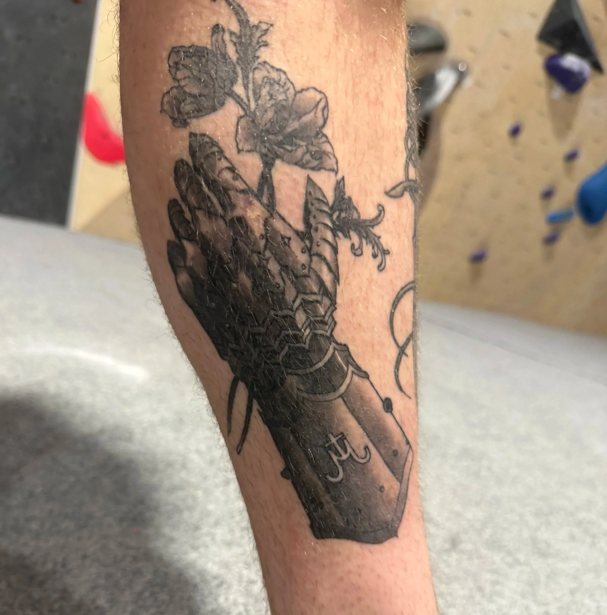 Black and gray tattoo of a flower being held by a hand, with a large knife or dagger below, on a person's forearm.
