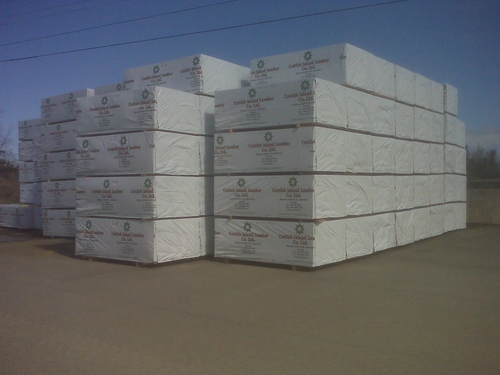 Stacks of white lumber wrapped in plastic with a label, placed outdoors on pavement under a blue sky.