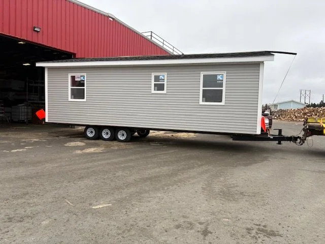 A tiny gray modular home on a trailer parked outside a red warehouse.