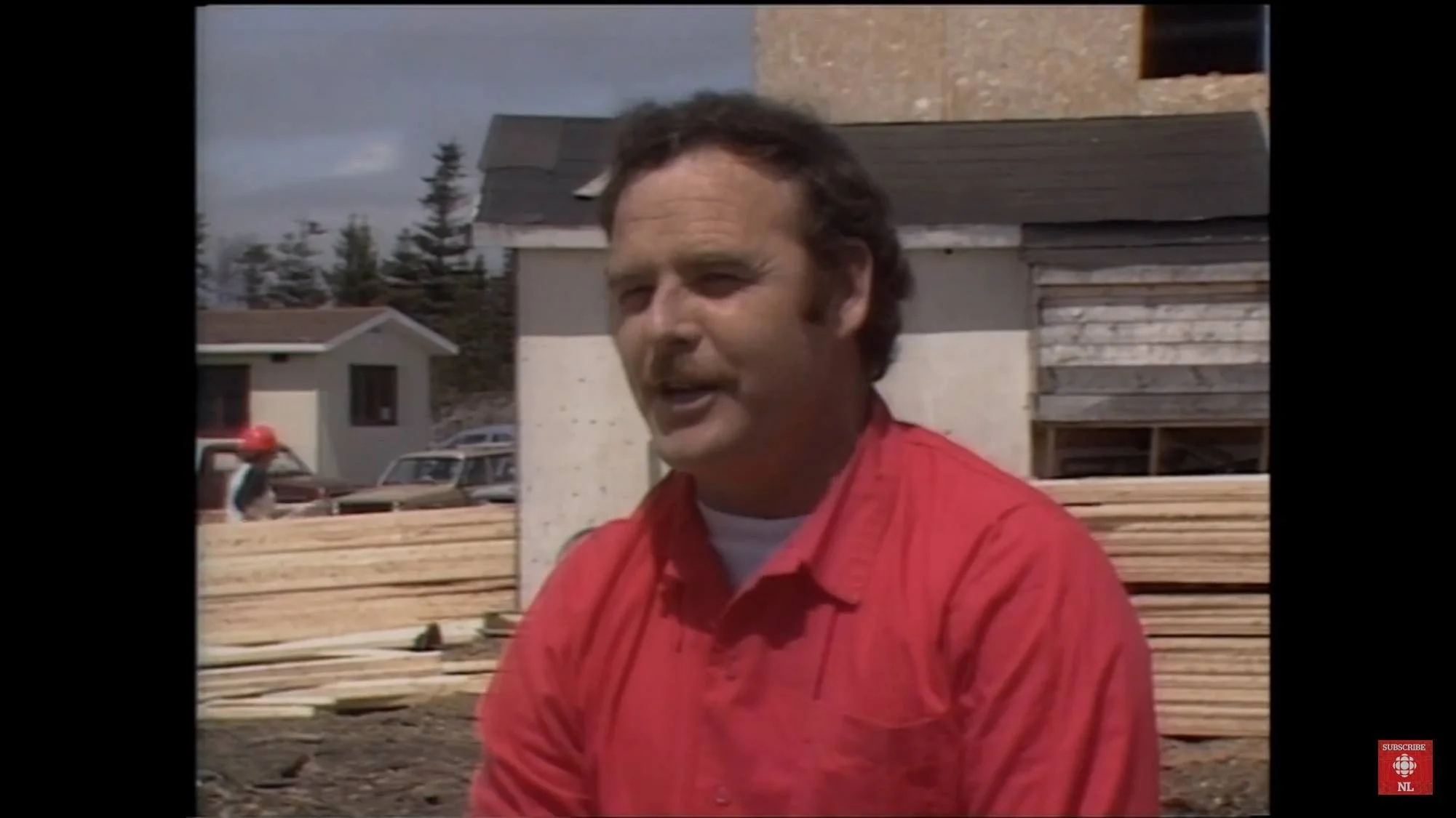 Rex Philpott, CEO of Cottle's Prefab Homes, is seen with dark hair and a mustache wearing a red shirt, standing outdoors in front of a construction site with stacked lumber. In the background, houses and parked cars are visible under a cloudy sky.