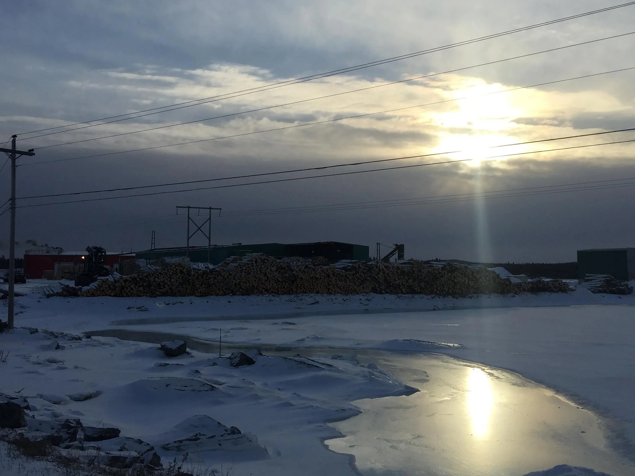 Snow-covered landscape with a Mill and Prefabricated Housing Facility on a frozen river, utility poles, logs stacked in the background, and a cloudy sky with the sun partially obscured.