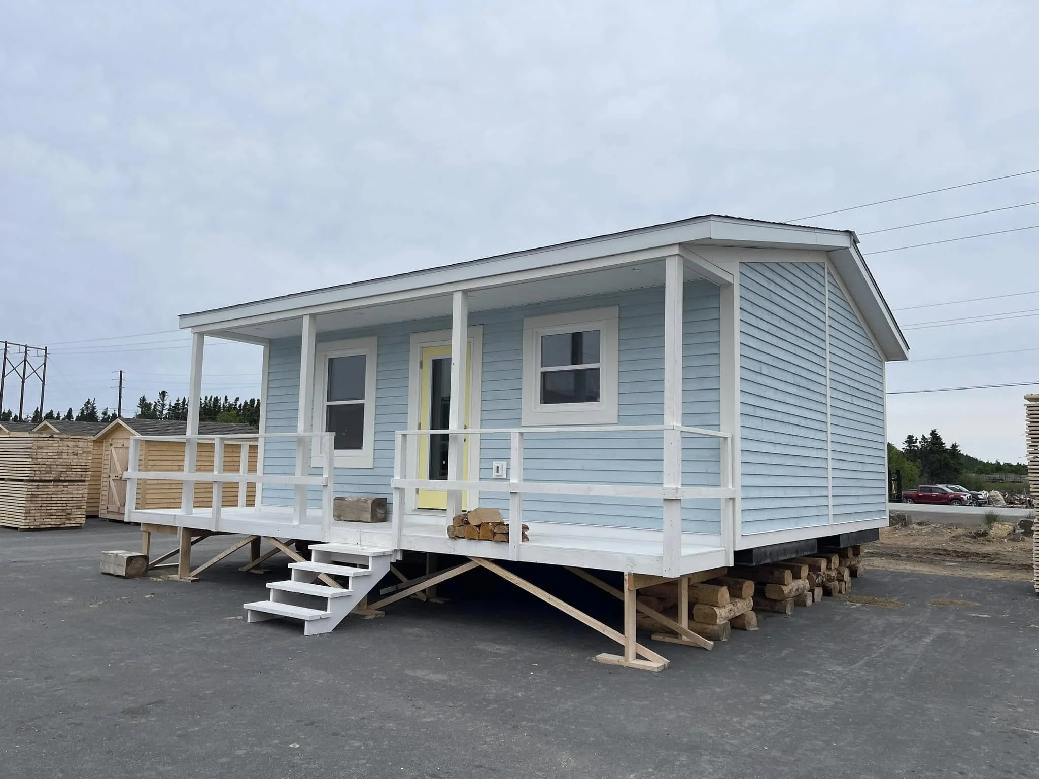 A blue modular home with a front porch, white trim, and yellow door, built on stilts with wood logs underneath, on a paved lot with other buildings and parked cars in the background.