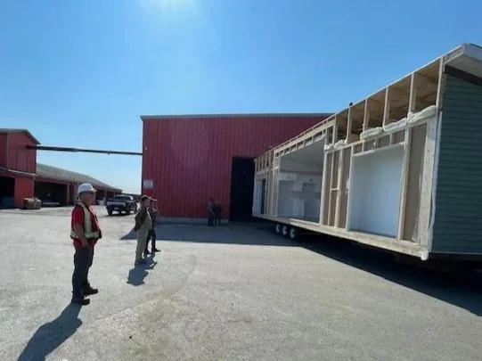 Construction workers at a building site with a partially built modular home on a truck outside a large red warehouse under a clear blue sky.