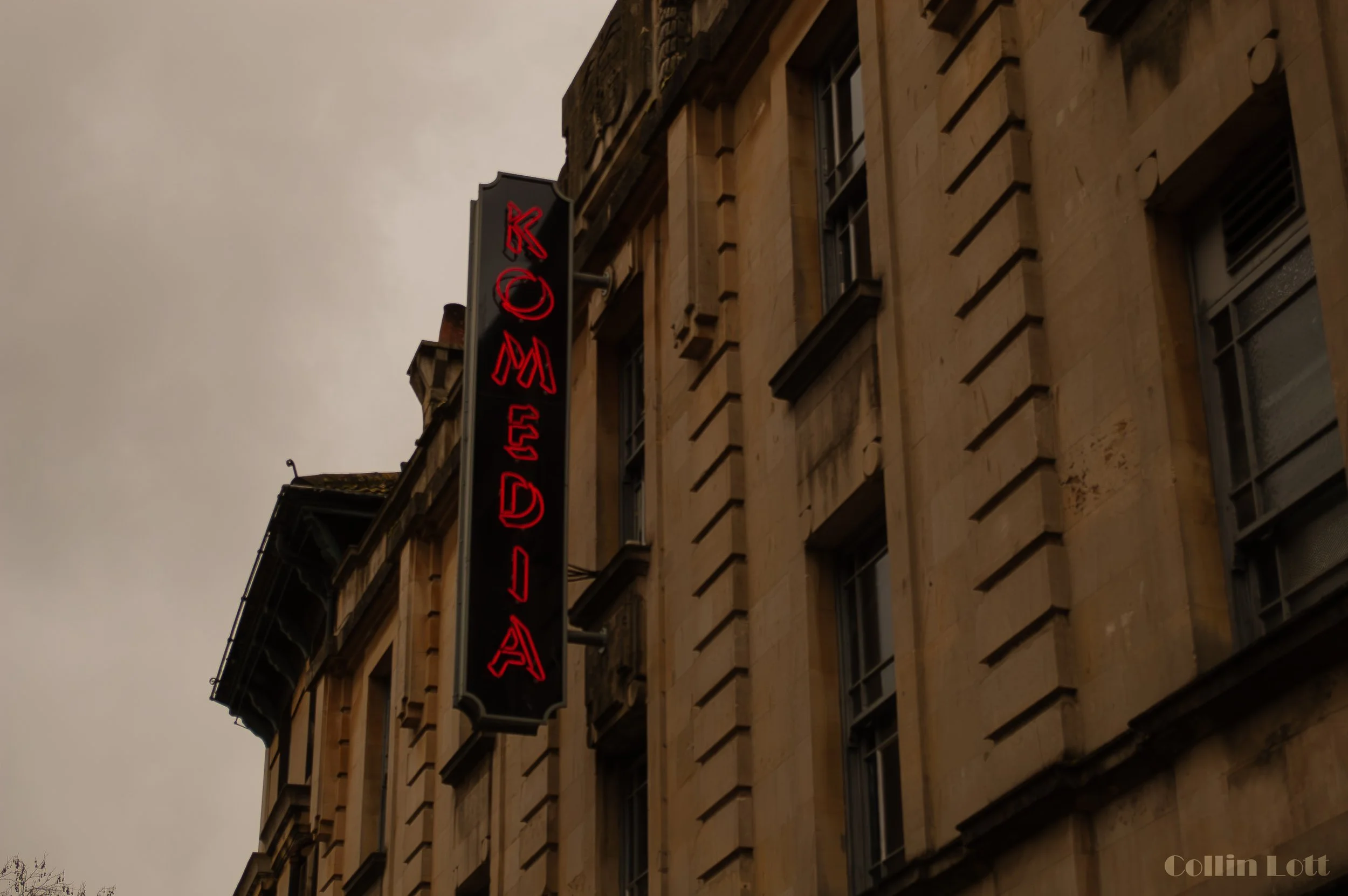 Neon sign with red lettering on black background attached to a beige stone building, with cloudy sky in the background.