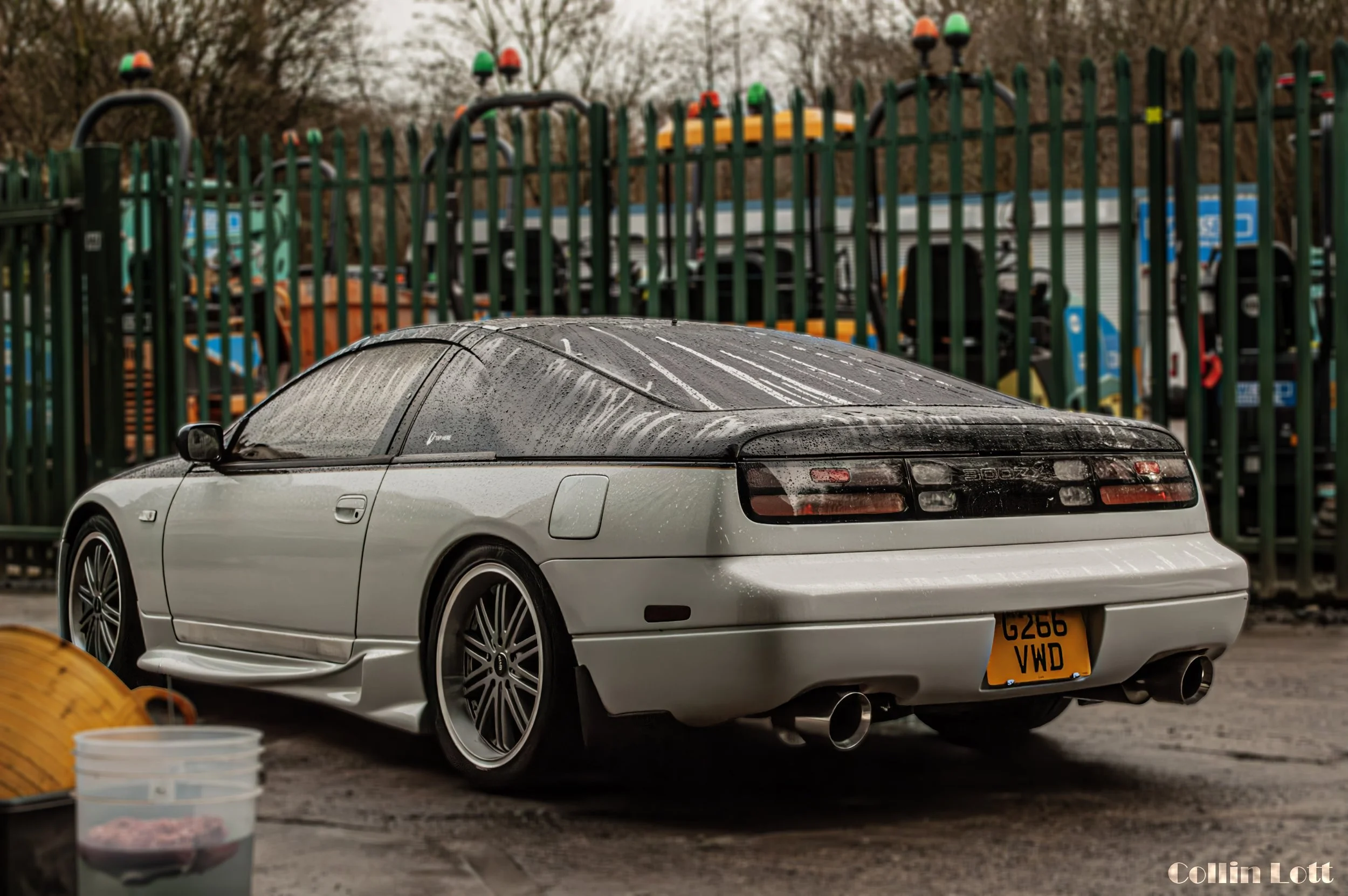 A silver sports car with black roof and tinted windows parked in a lot at a car wash, with water droplets on the surface and a yellow license plate reading G266 VWD. There is a green fence and equipment in the background, and some buckets and cleaning supplies in the foreground.