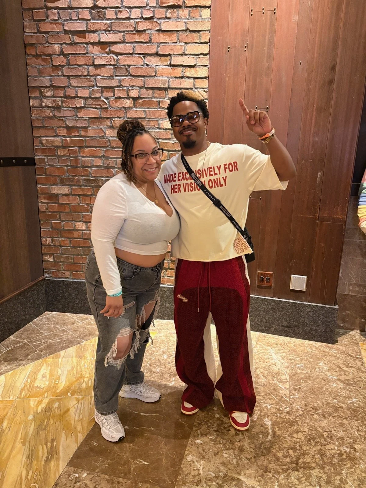 Two people standing together indoors against a brick wall and wooden paneling, smiling at the camera. The woman is wearing glasses, a white long-sleeve crop top, ripped jeans, and white sneakers. The man is wearing sunglasses, a cream-colored T-shirt