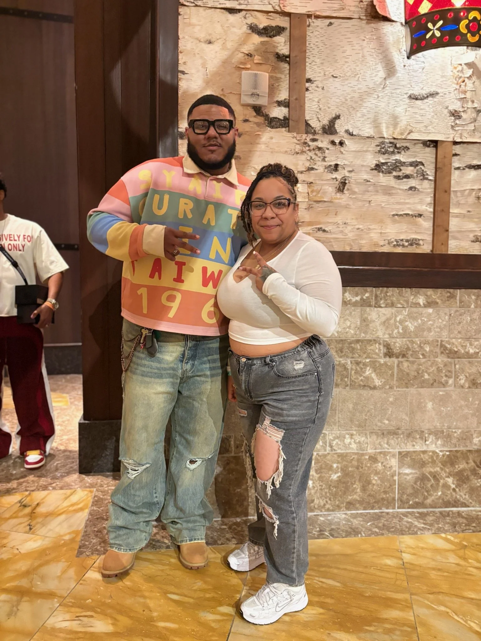 Two people standing indoors, posing and making peace signs. The person on the left is a tall man with glasses, wearing a colorful long-sleeve shirt and ripped jeans. The person on the right is a woman with glasses, wearing a white long-sleeve top and