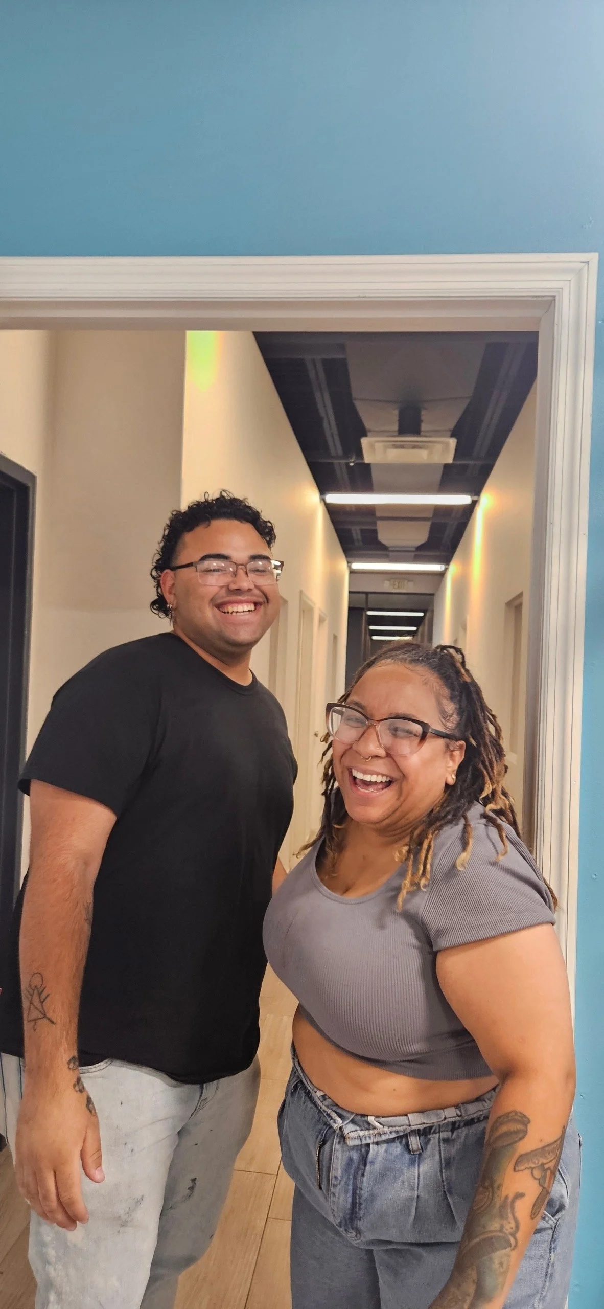 Two smiling women and a man standing together in a hallway, laughing and posing for a photo.