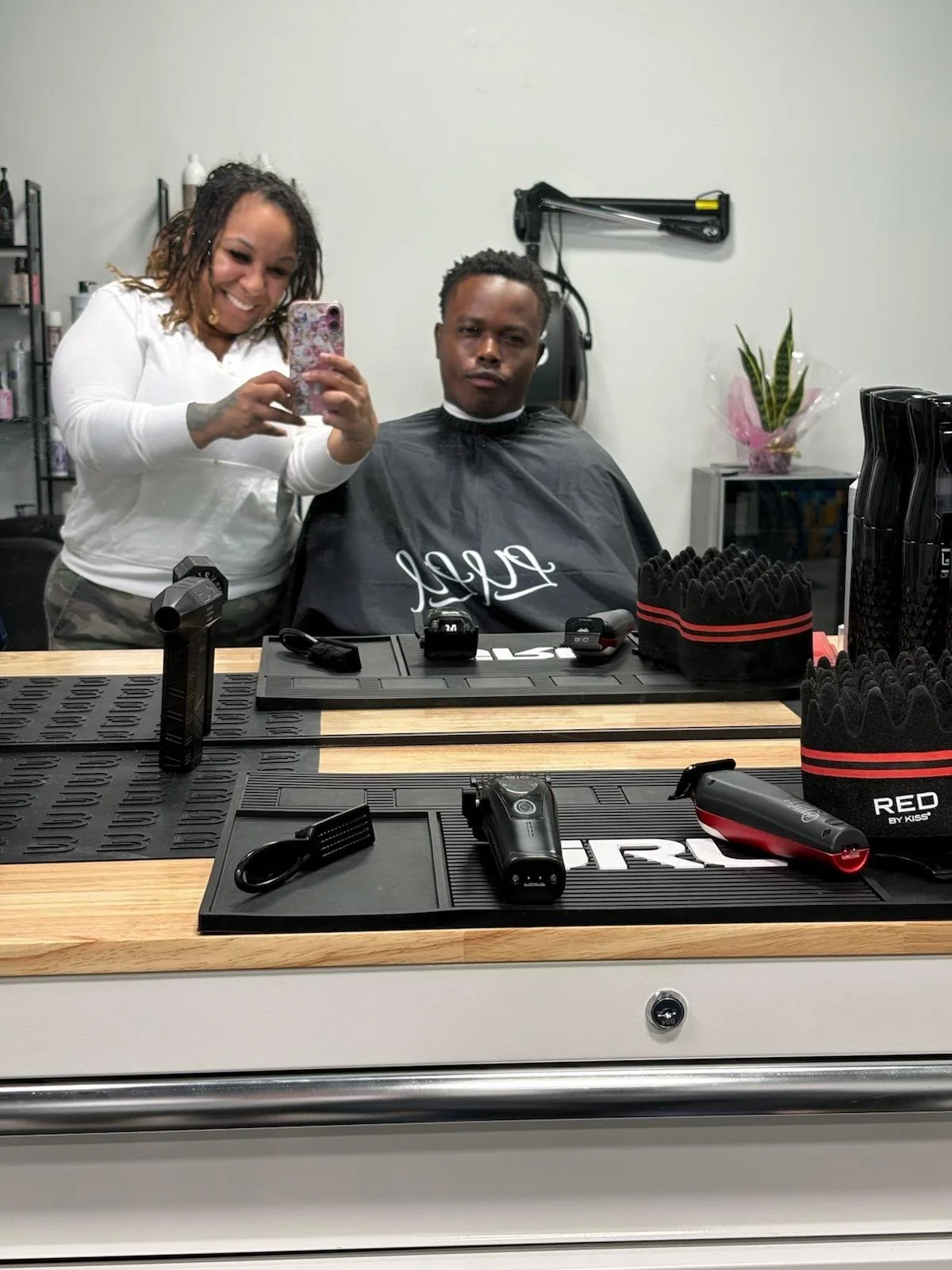 A woman is taking a selfie with a man in a barber's chair at a barbershop, with grooming tools and products on the counter in front of them.
