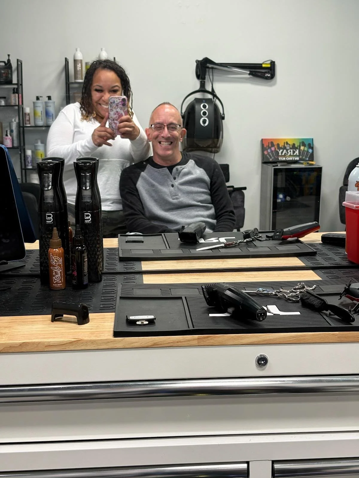 A woman taking a selfie with a man sitting in a barber's chair in a barber shop. The countertop has hair styling tools, sprays, and collectibles, with shelves and a wall-mounted vacuum in the background.