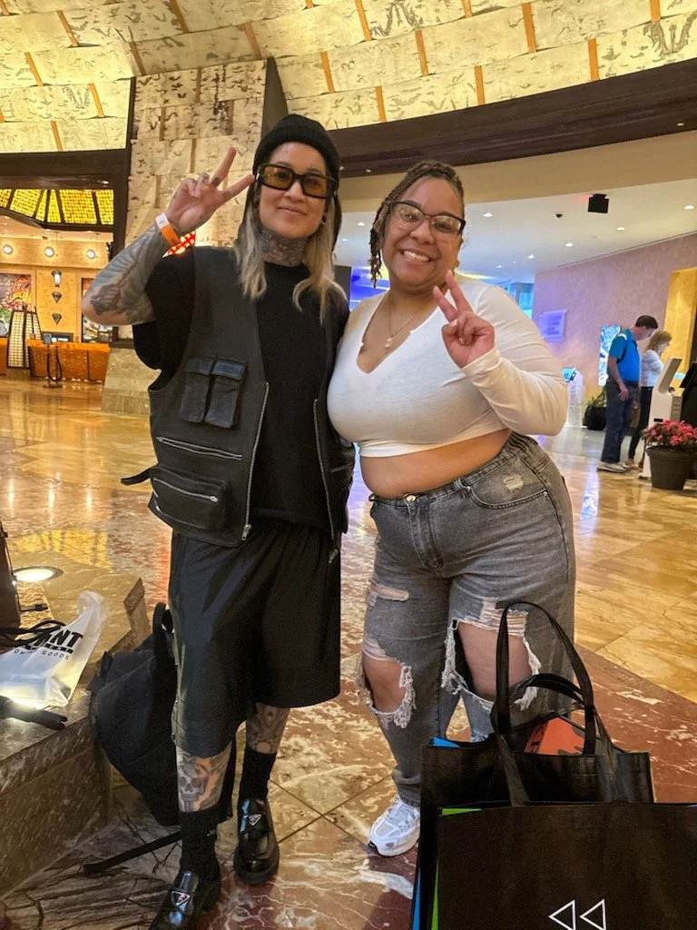 Two women smiling and making peace signs pose together in a lobby area. One woman is wearing a black beanie, glasses, a black jacket with multiple pockets, black shorts, and black shoes, with tattoos visible on her legs. The other woman is wearing gl