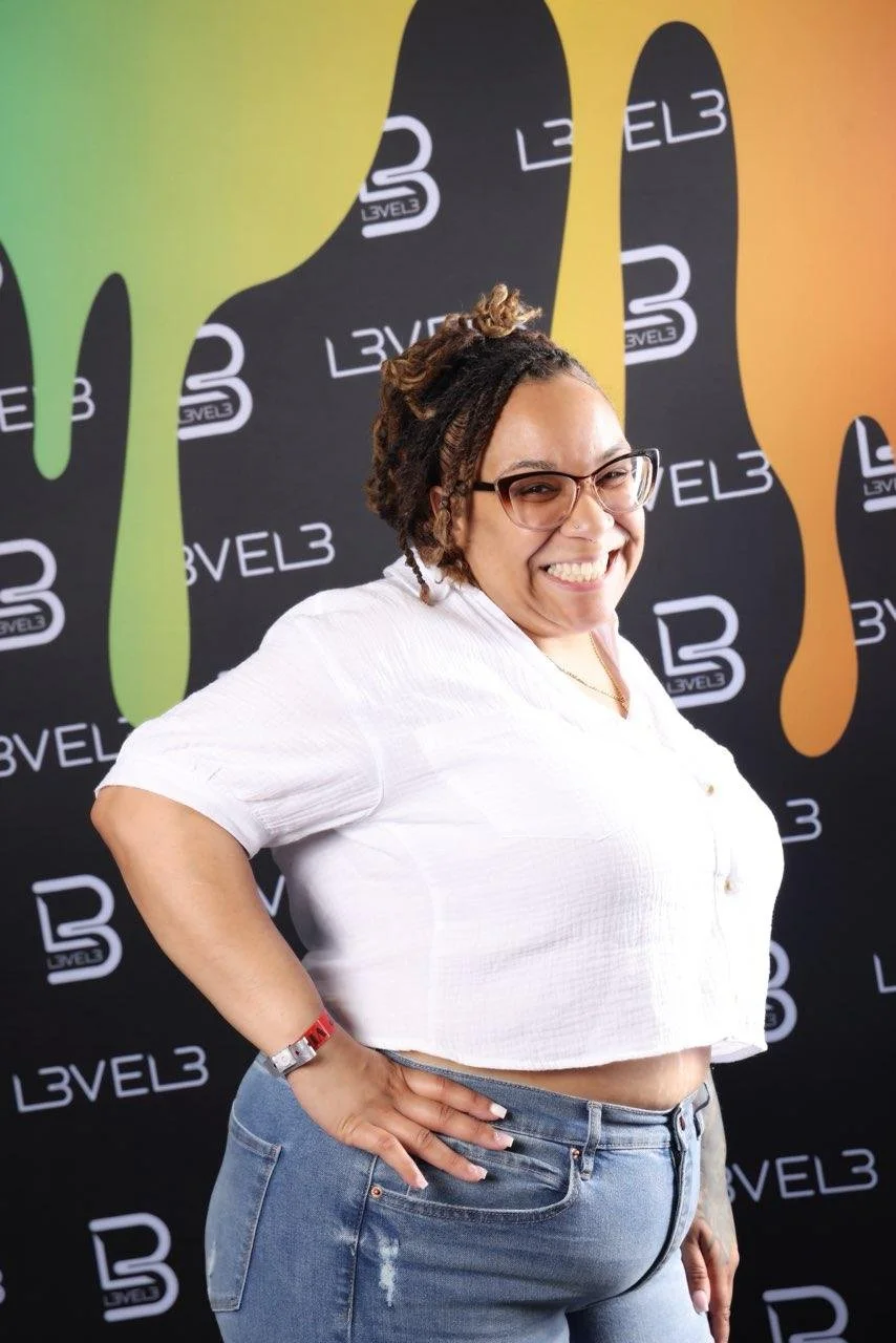 A smiling woman with glasses, wearing a white cropped shirt and jeans, standing in front of a black backdrop with colorful abstract shapes and the text 'LEVEL3' printed repeatedly.