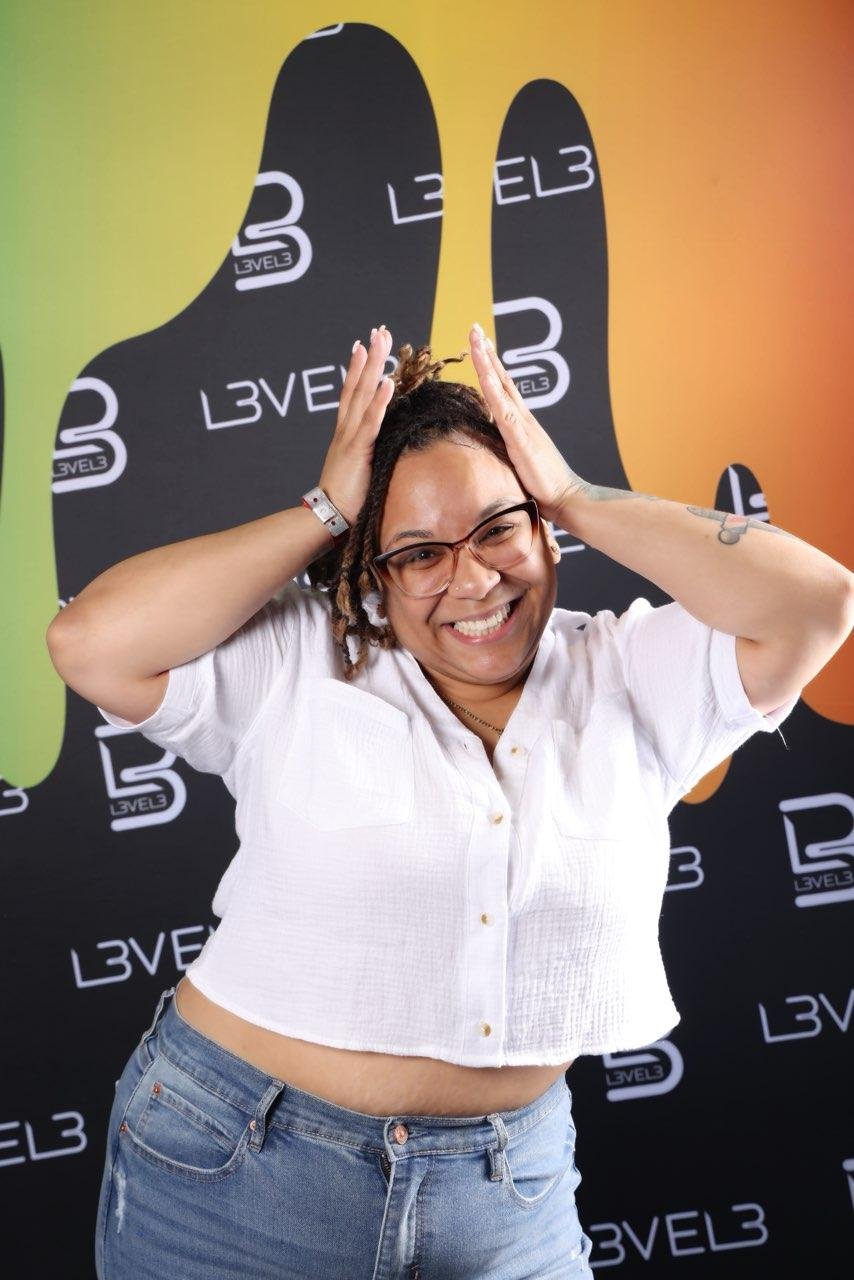 A woman with glasses and dreadlocks smiling and holding her hands on her head in front of a colorful backdrop with the logo L3VEL3.