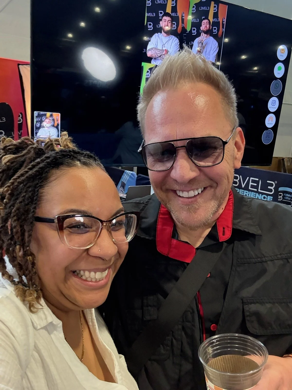 Two people smiling for a selfie in front of a large screen at an event. The woman on the left has glasses, a nose piercing, and dreadlocks. The man on the right has sunglasses, short blond hair, and is holding a drink.