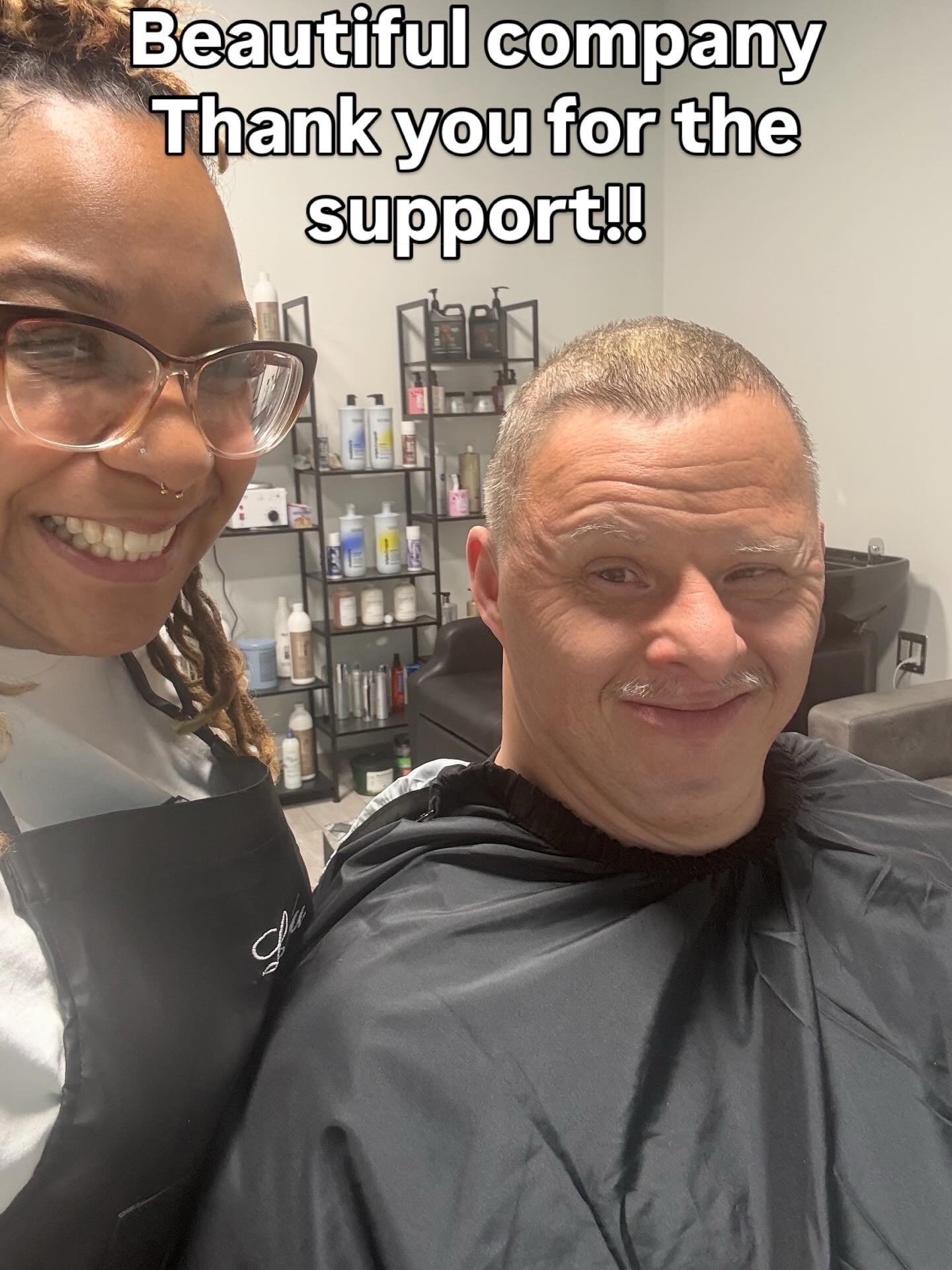 A woman and a man inside a hair salon, smiling; the woman is wearing glasses and a black apron, and the man is wearing a black haircut cape; shelves with hair products are visible in the background; text overlay says 'Beautiful company. Thank you for