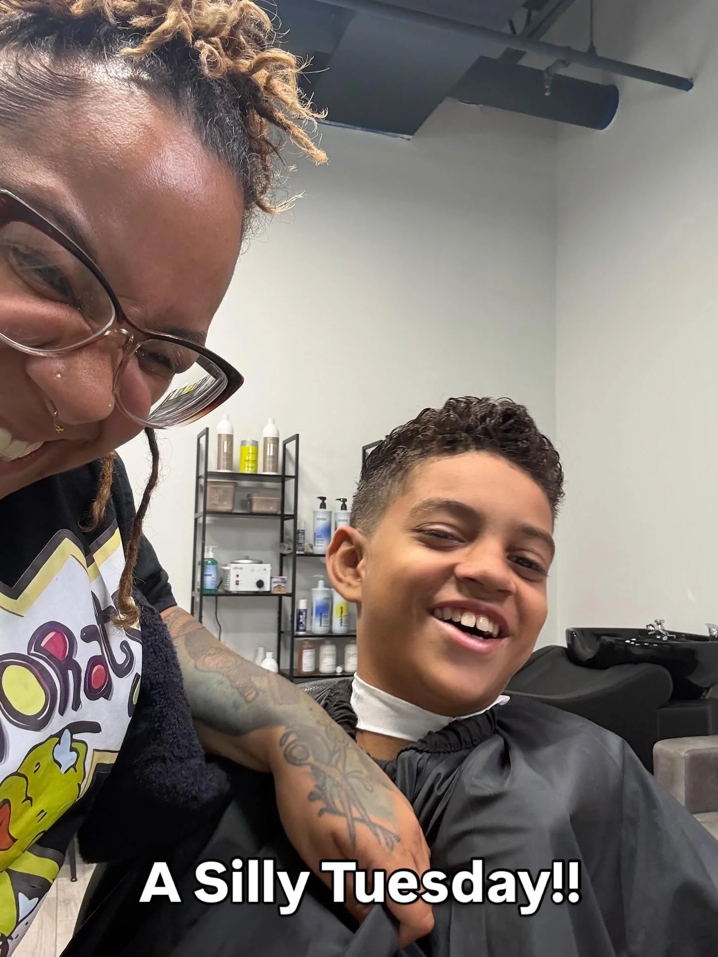 A woman with glasses and tattoos on her arm is giving a boy a haircut. They are smiling and appear to be having fun. The setting is a hair salon with shelves of hair products in the background.