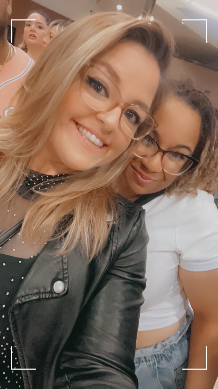 Two women wearing glasses taking a selfie together at an indoor event, smiling and standing close to each other.
