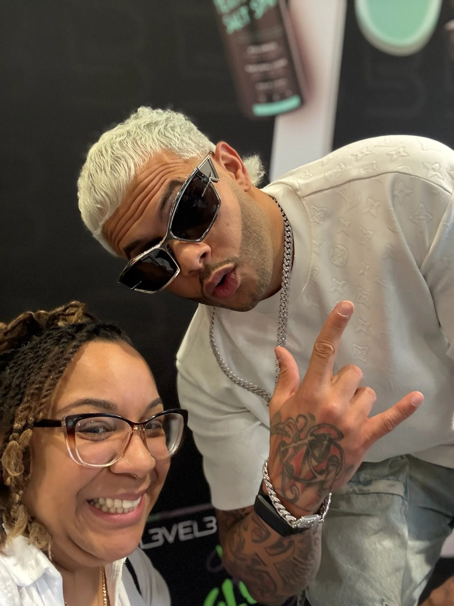 A woman with glasses smiling and a man with platinum blonde hair, sunglasses, and tattoos making a rock hand gesture.