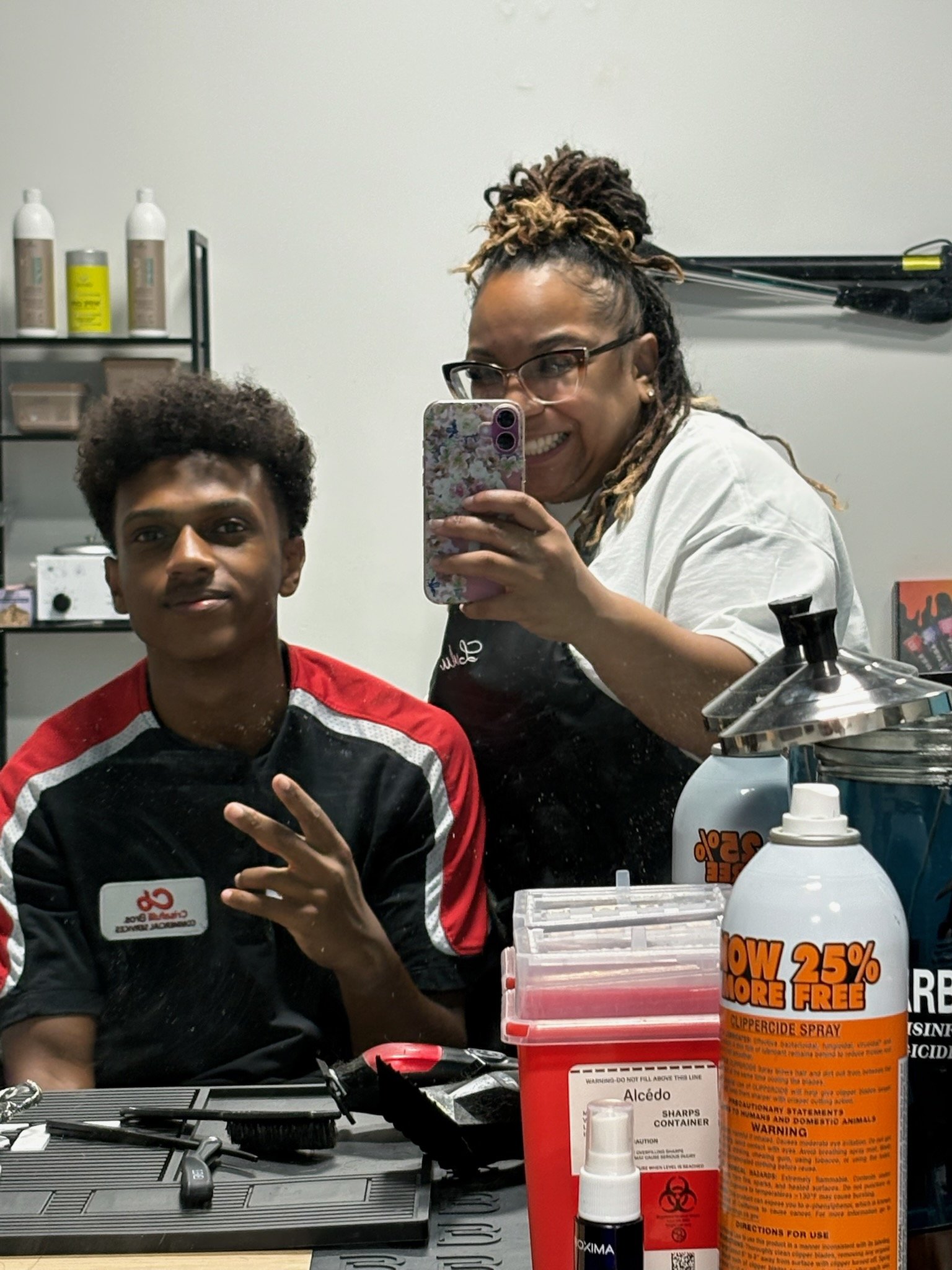 Two people taking a mirror selfie in a salon, with hair and beauty products on the table and shelves. One person is sitting, the other is standing and holding a phone. They are smiling.