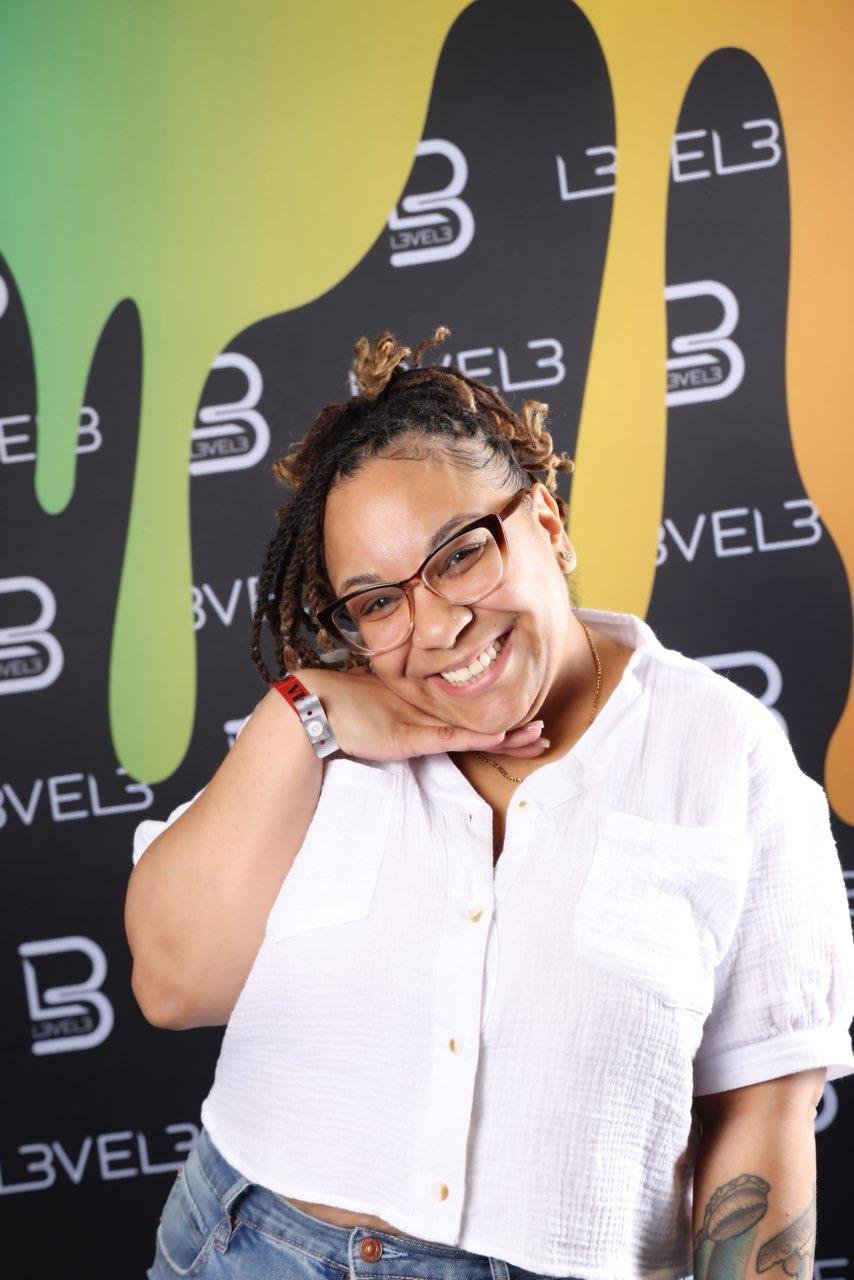 Smiling woman with glasses and dreadlocks in front of a colorful background with the words 'LEVEL 3' repeated.