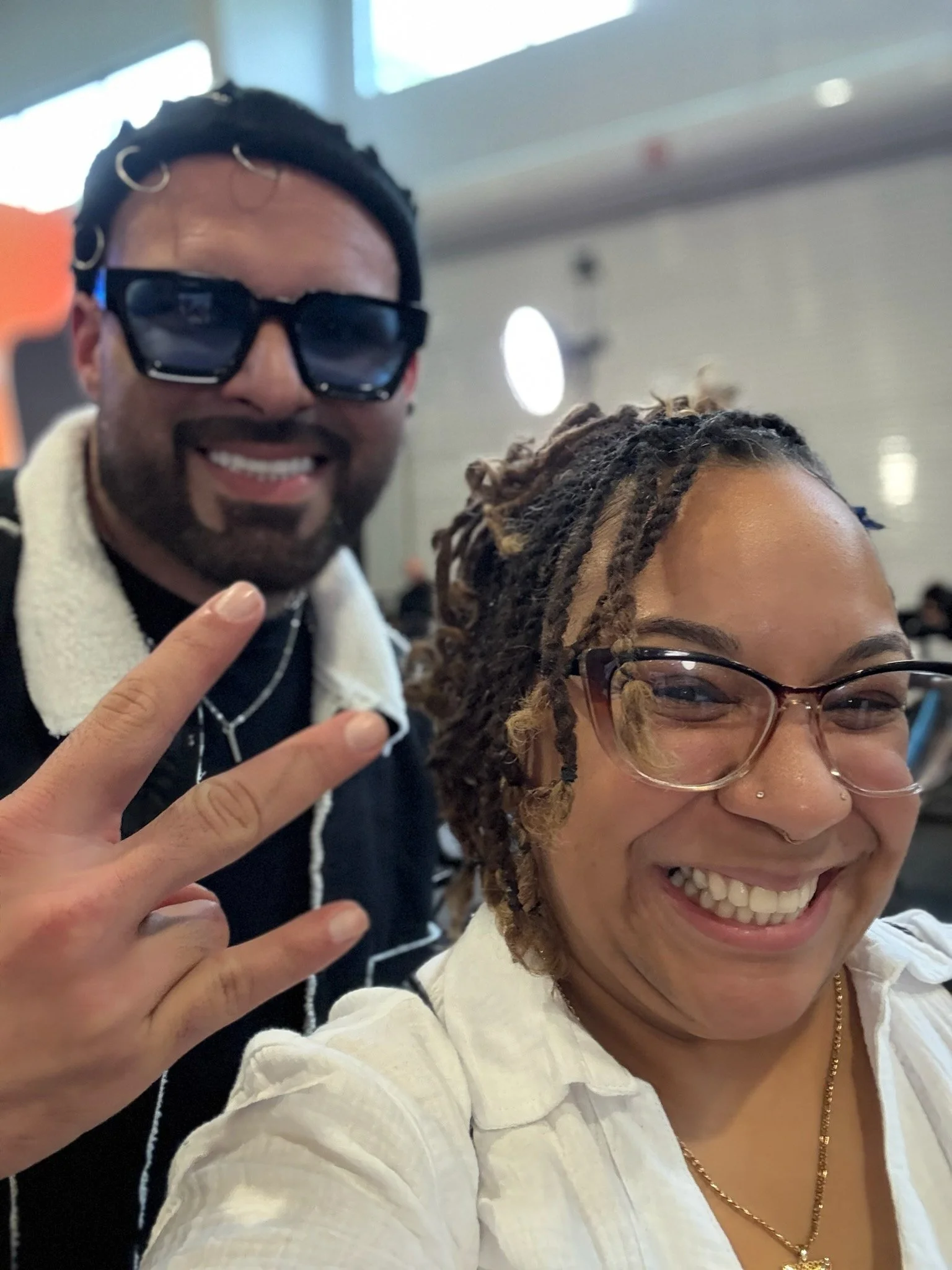 A man and woman smiling for a selfie indoors. The man, wearing large sunglasses and making a peace sign, has dark curly hair with rings, a beard, and a mustache. The woman, with glasses, dreadlocks, and a nose ring, is smiling broadly, wearing a whit