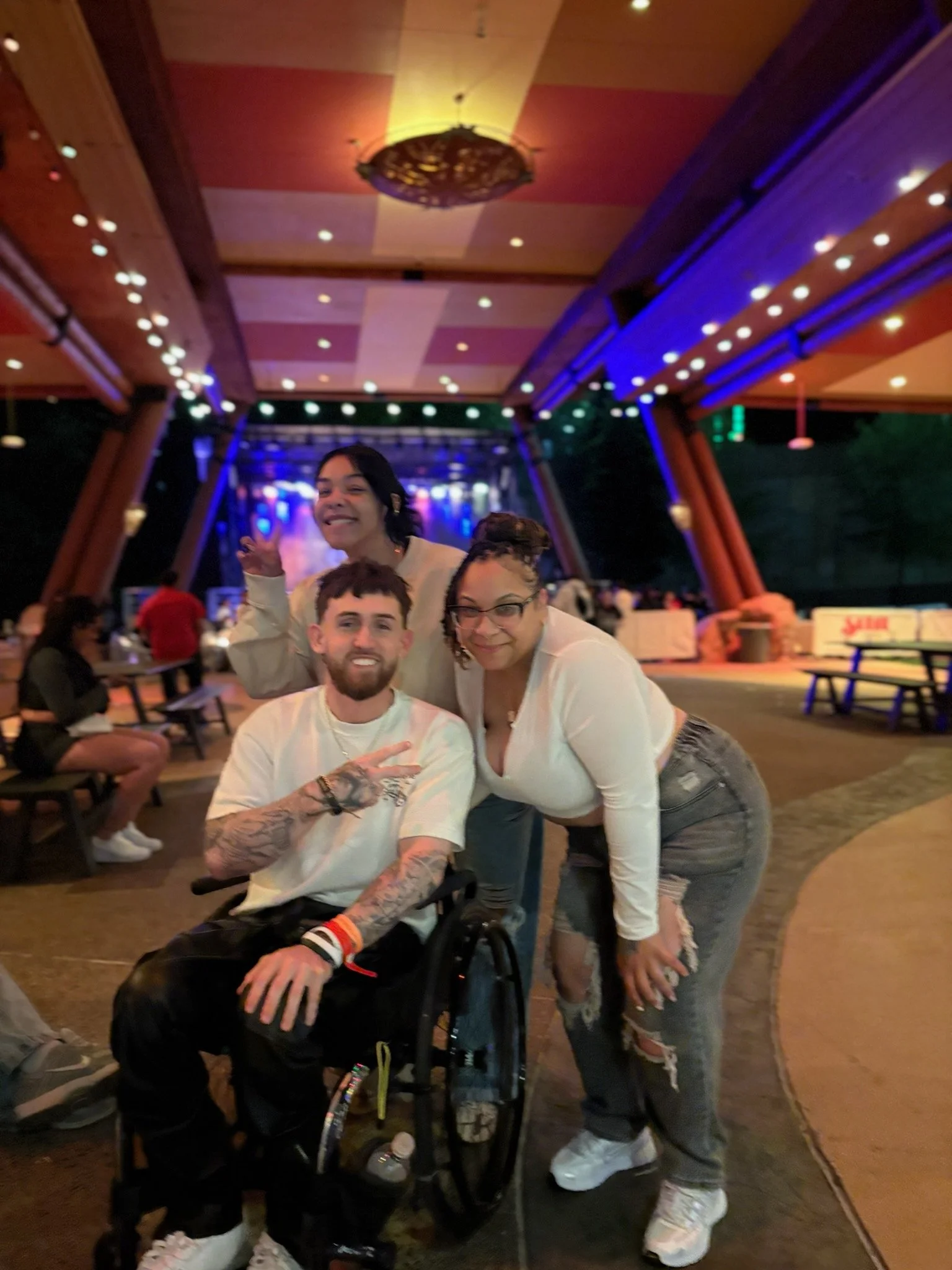 Three smiling young adults posing together in a night club or concert venue with colorful lighting, one seated in a wheelchair and two standing, all dressed casually.