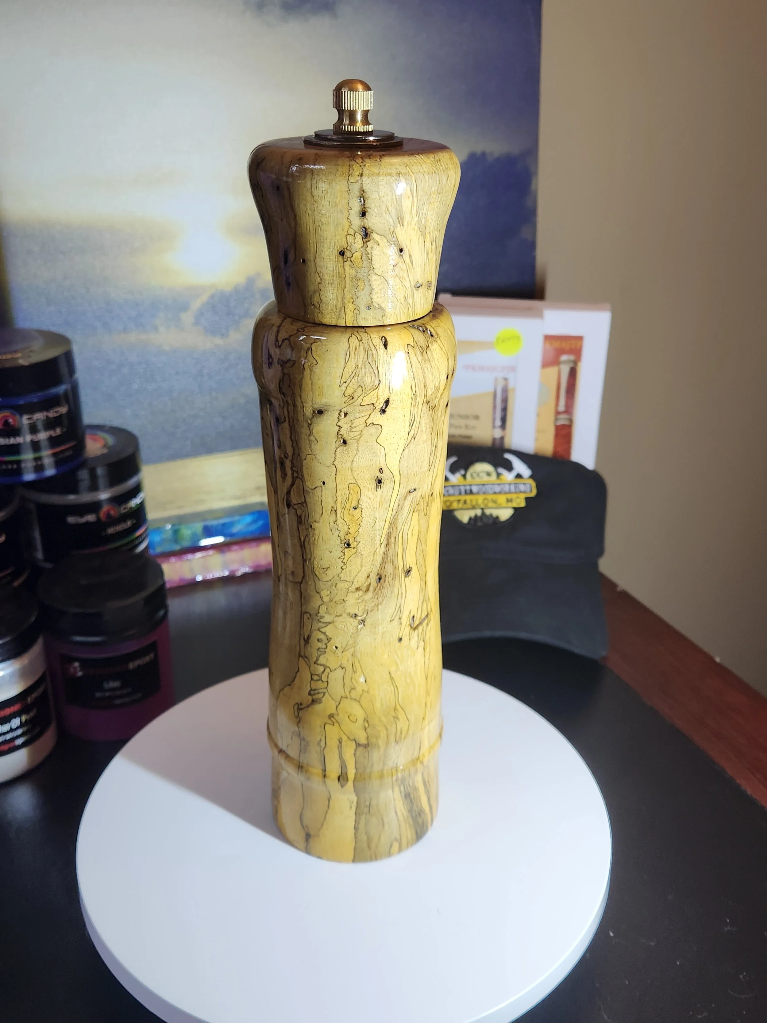 Wooden pepper mill on a white circular base, with a background of various objects including a black cap, jars, and boxes.