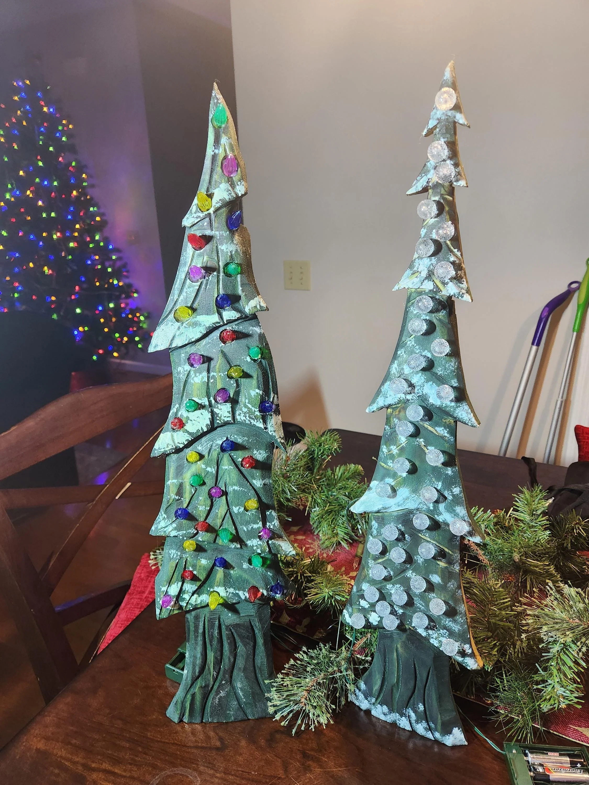 Decorative Christmas trees with colorful ornaments and dots, placed on a table with greenery, with a decorated Christmas tree in the background.