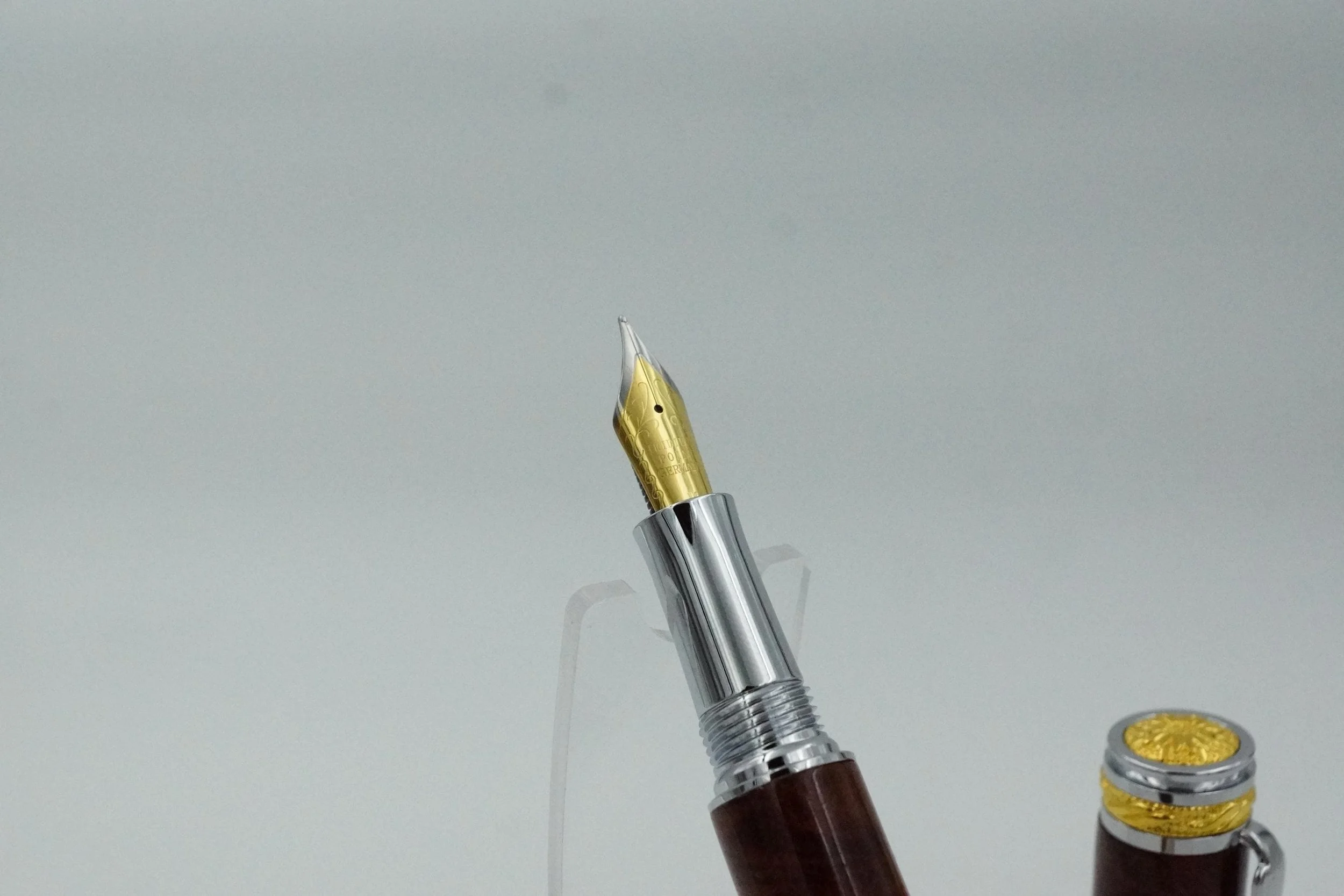 Close-up of a fountain pen with a gold nib and a dark brown body, with a golden decorative element on the cap, against a plain background.