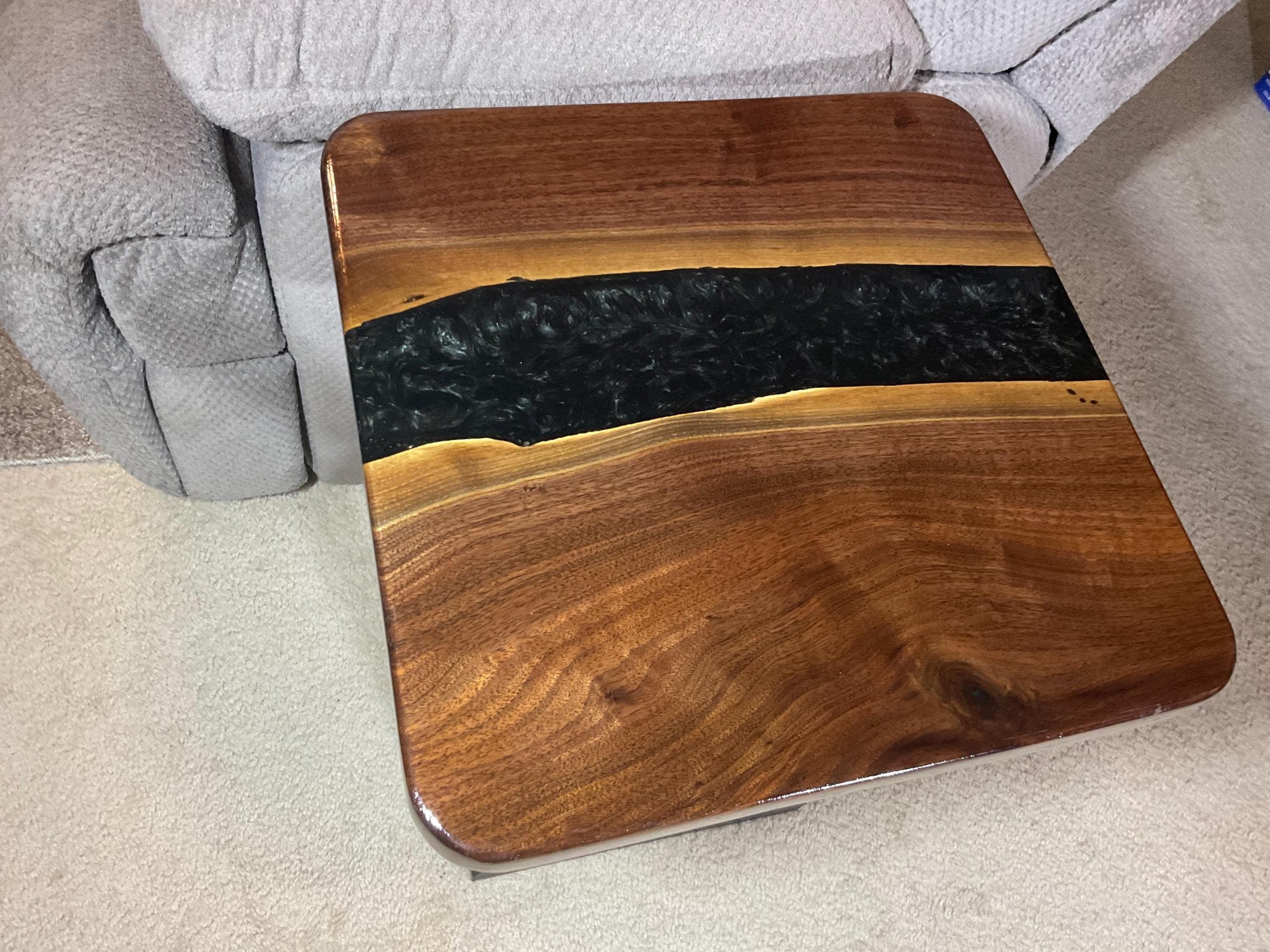 Square wooden and resin side table with a live edge design, placed next to a gray upholstered sofa, on a beige carpeted floor.