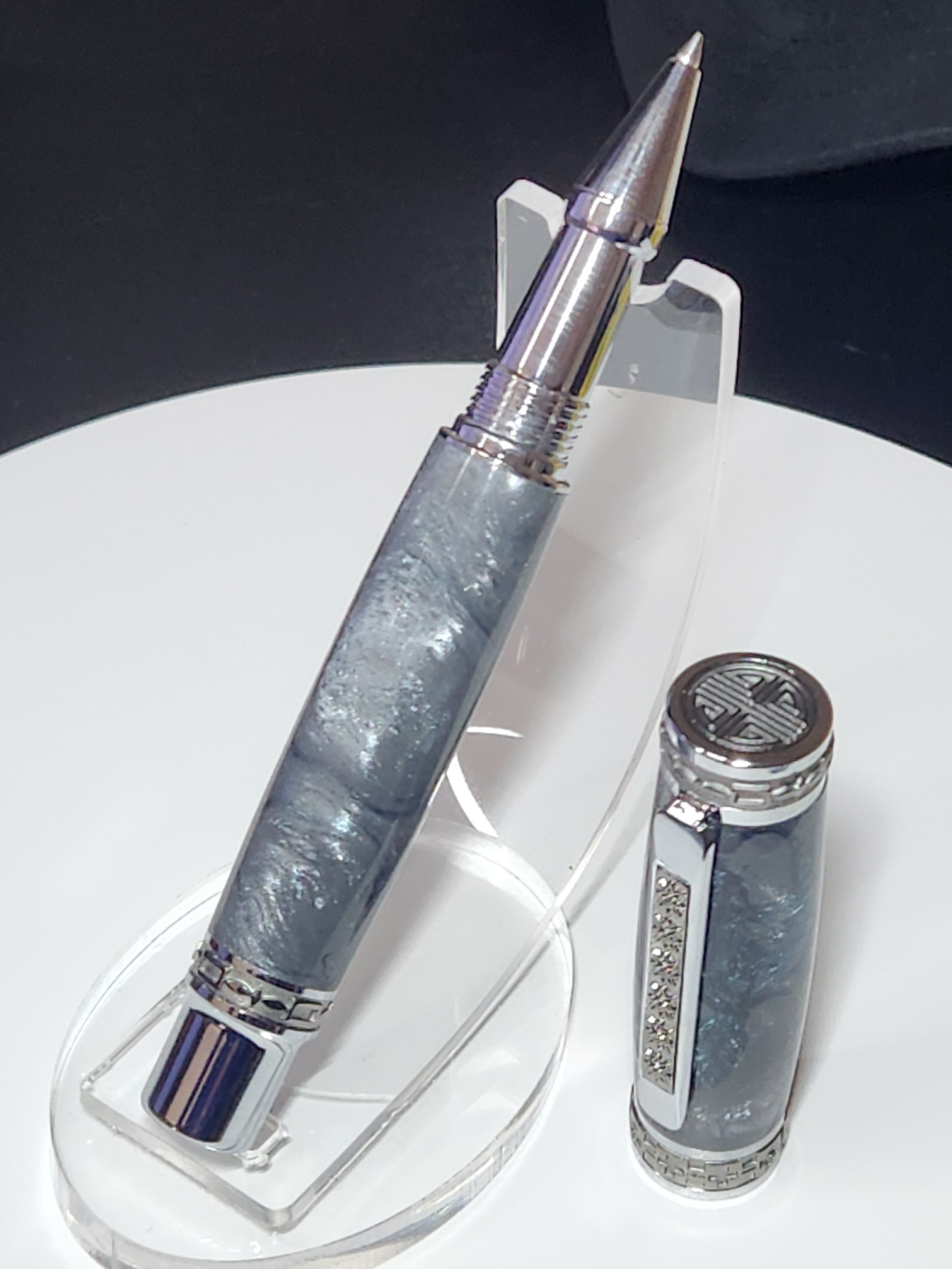 A silver and gray luxury ballpoint pen and a matching rollerball pen on a clear display stand on a white surface.