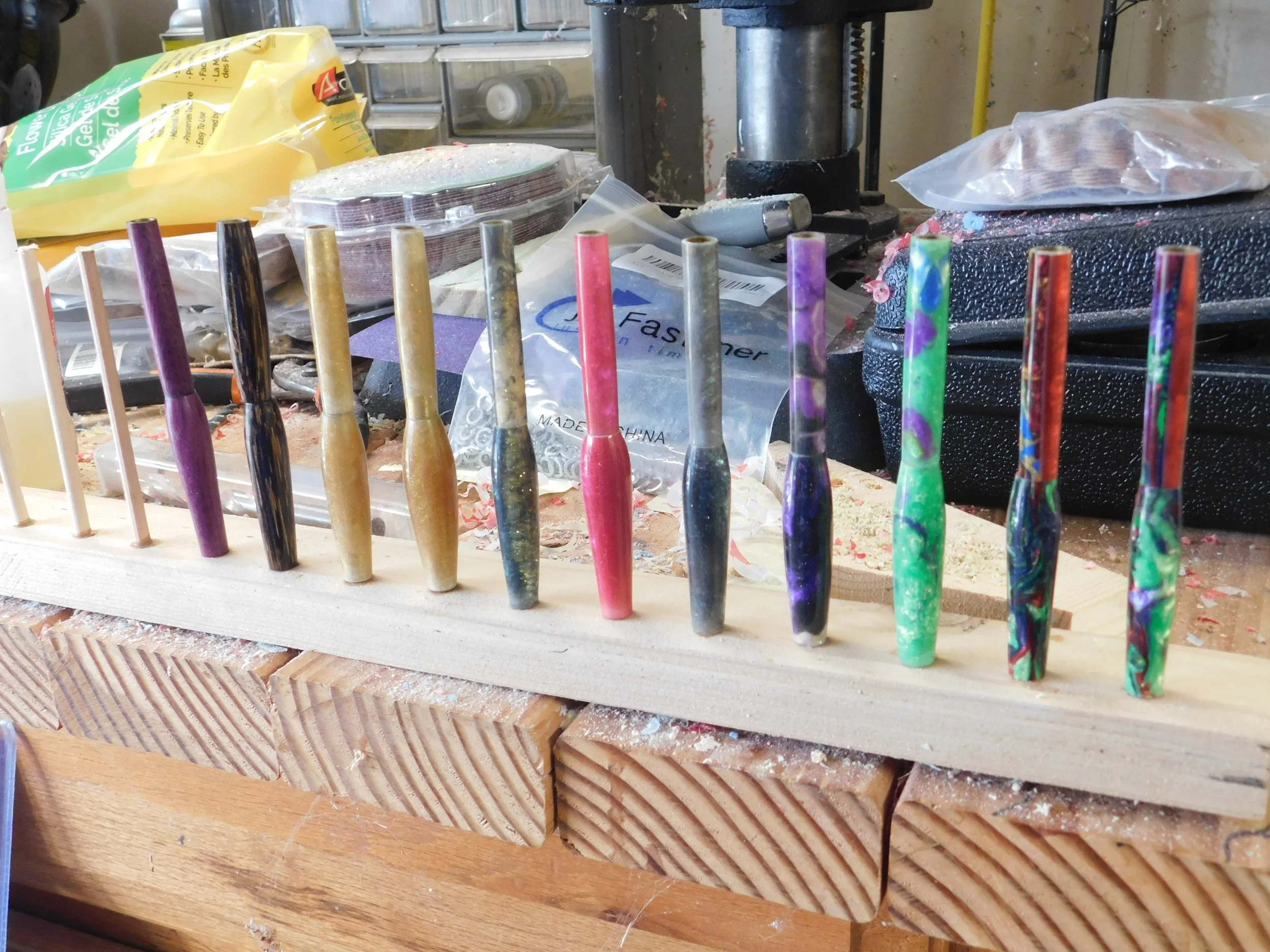 A wooden board with colorful resin pens in various swirling patterns, arranged in a line on a cluttered workbench with woodworking tools and supplies in the background.